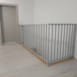 PREMIUMSTAL SEBASTIAN PRZYBYSZ - Szara, metalowa balustrada schodowa z pionowymi szczeblami, zabezpieczająca schody. Widok z boku, na jasnym tle ściany i podłogi. Minimalistyczny design.