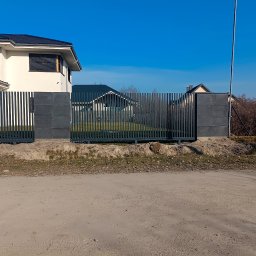 PREMIUMSTAL SEBASTIAN PRZYBYSZ - Nowoczesne ogrodzenie panelowe w kolorze grafitowym z betonowymi słupkami, na tle domu jednorodzinnego i błękitnego nieba. Widok z zewnątrz posesji.