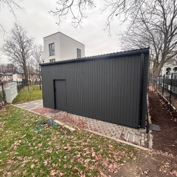 PREMIUMSTAL SEBASTIAN PRZYBYSZ - Ciemnoszara, pionowo panelowa konstrukcja z drzwiami, obok czarny panelowy płot i trawnik z liśćmi. W tle budynek mieszkalny i drzewa bez liści.