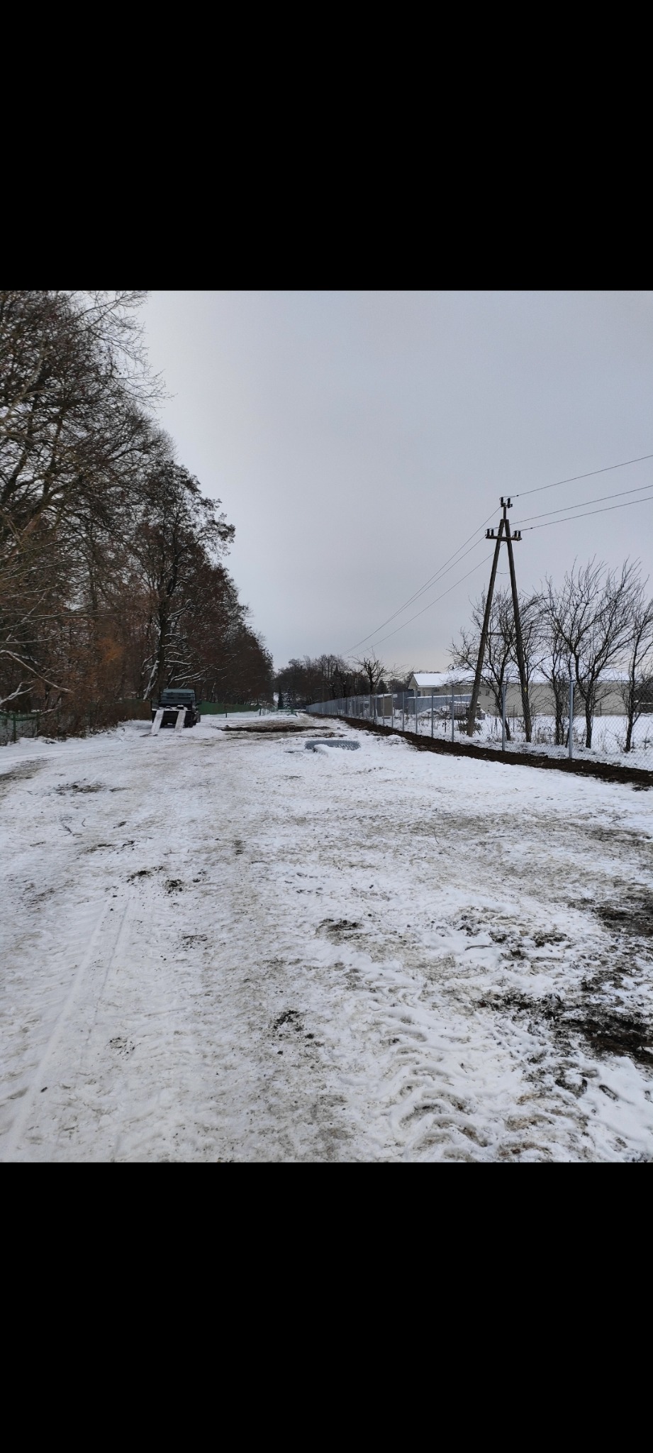 Zimowy krajobraz z widokiem na nowo postawione ogrodzenie z siatki, częściowo zasypane śniegiem, z widocznym autem dostawczym w tle i słupami energetycznymi