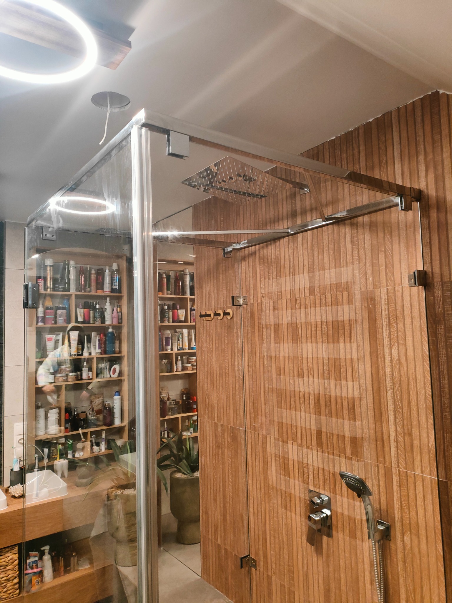 Szklana kabina prysznicowa z chromowanymi okuciami i deszczownicą, drewnopodobne panele ścienne, półki z kosmetykami widoczne przez szkło.