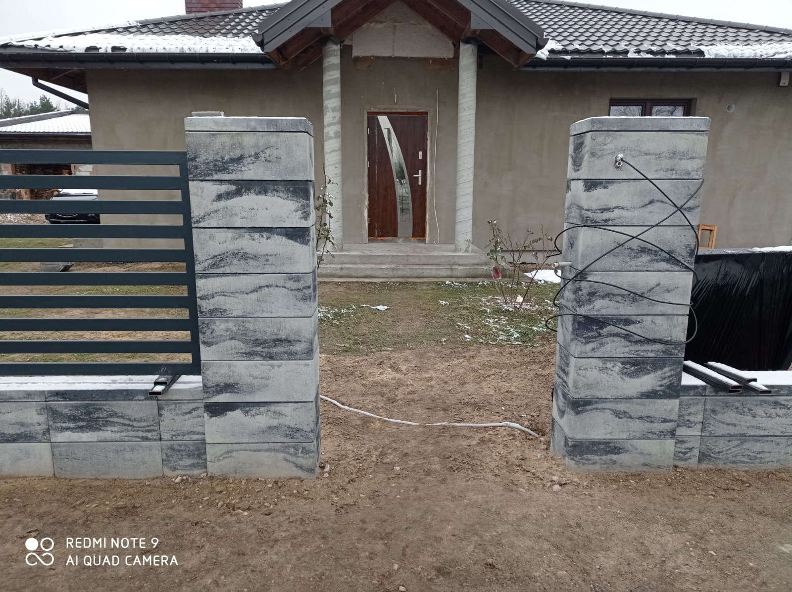 Fragment nowo postawionego ogrodzenia z paneli metalowych i słupków betonowych o szarej barwie, widok na dom w trakcie wykończenia, kabel elektryczny prowizorycznie przymocowany do słupka.