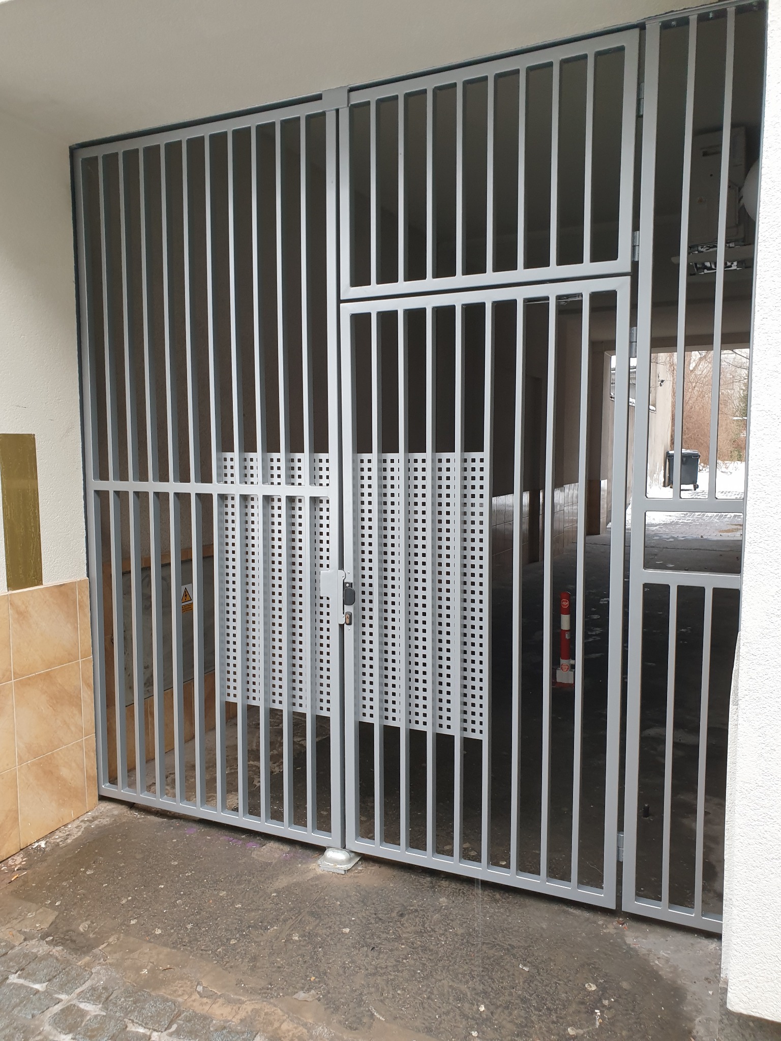 Szara, metalowa brama wjazdowa z pionowych prętów, częściowo otwarta, z widoczną kratką wentylacyjną w tle, zamontowana przy wejściu do garażu.