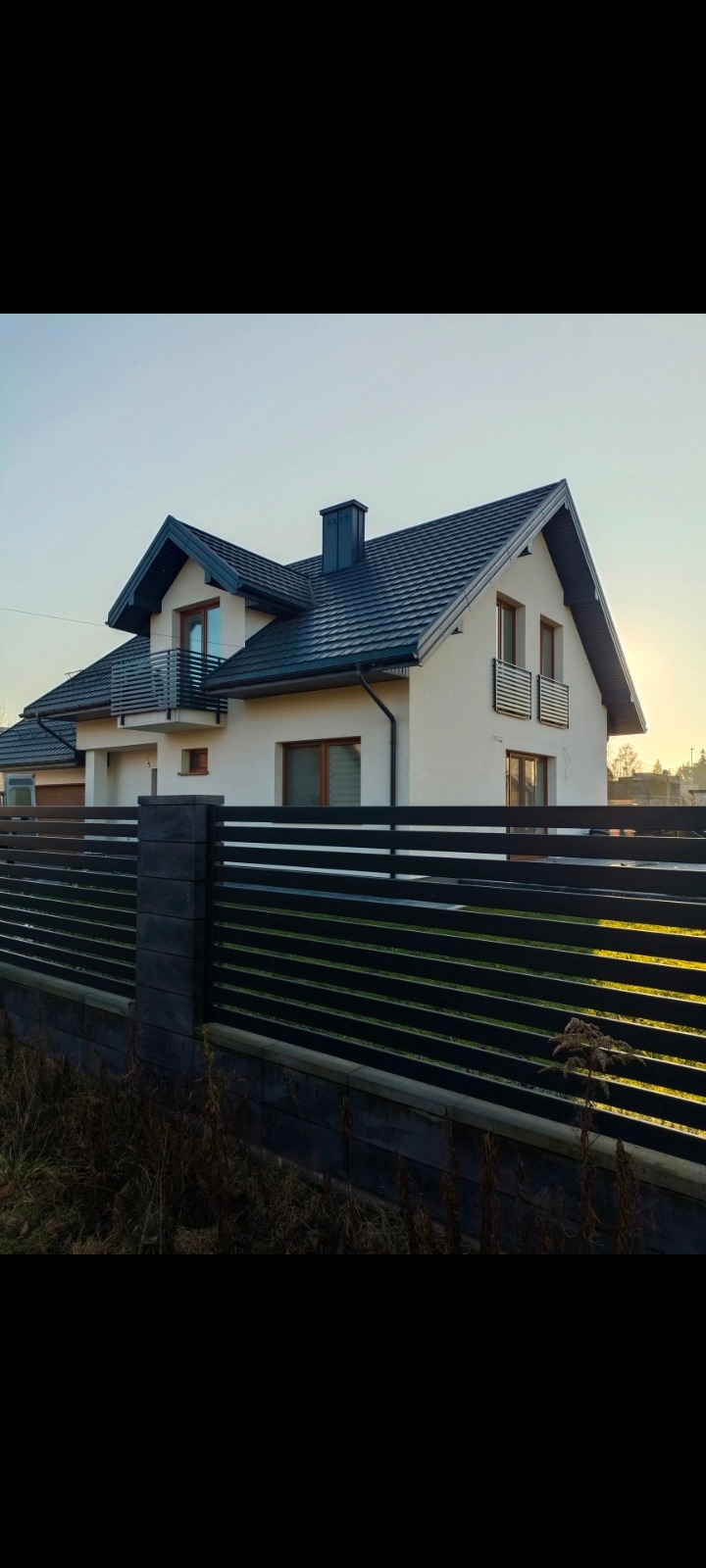 Nowoczesny dom z czarnym, poziomym ogrodzeniem panelowym na tle błękitnego nieba. Widoczne detale: betonowe słupki i podmurówka. Ujęcie z poziomu gruntu.