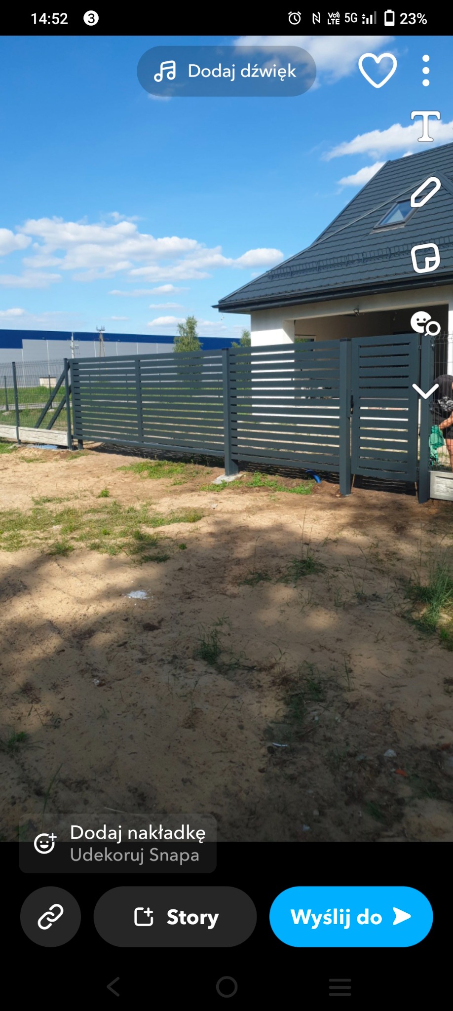 Nowoczesne, poziome ogrodzenie panelowe w kolorze grafitowym, z furtką, na tle domu z szarą dachówką i błękitnego nieba. Widoczne słupki i przęsła ogrodzenia.