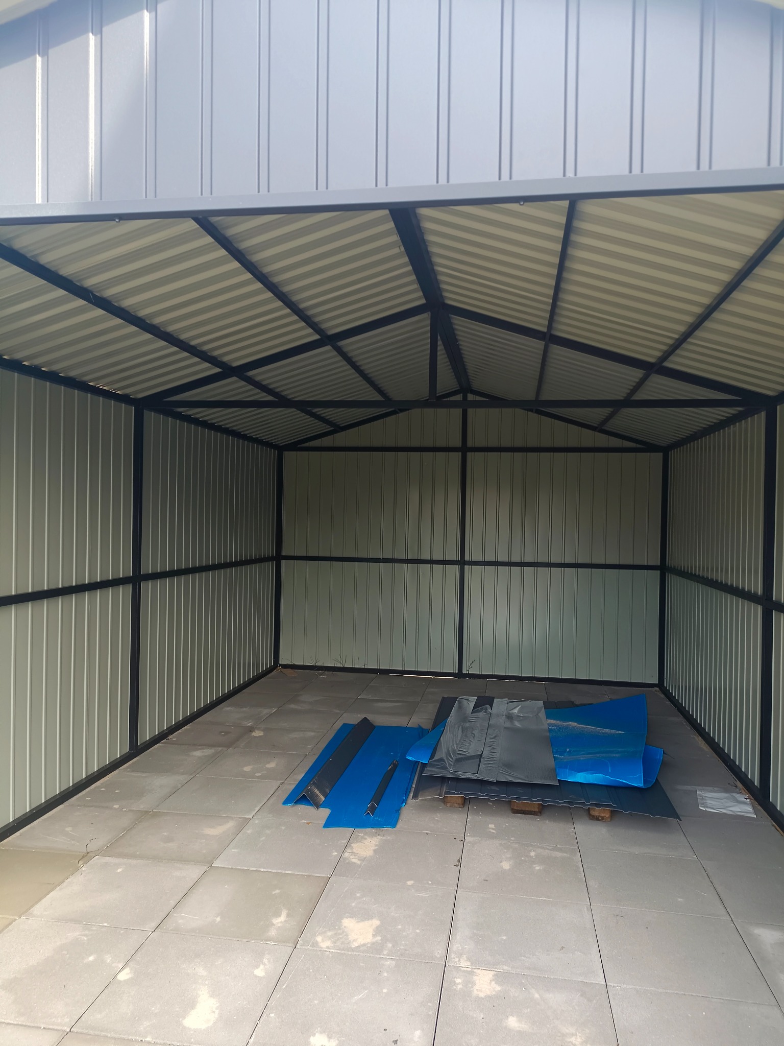 Metalowa konstrukcja garażu z blachy falistej, widok od wewnątrz. Na podłodze leżą arkusze blachy w kolorze niebieskim i szarym. Widoczne łączenia spawane.