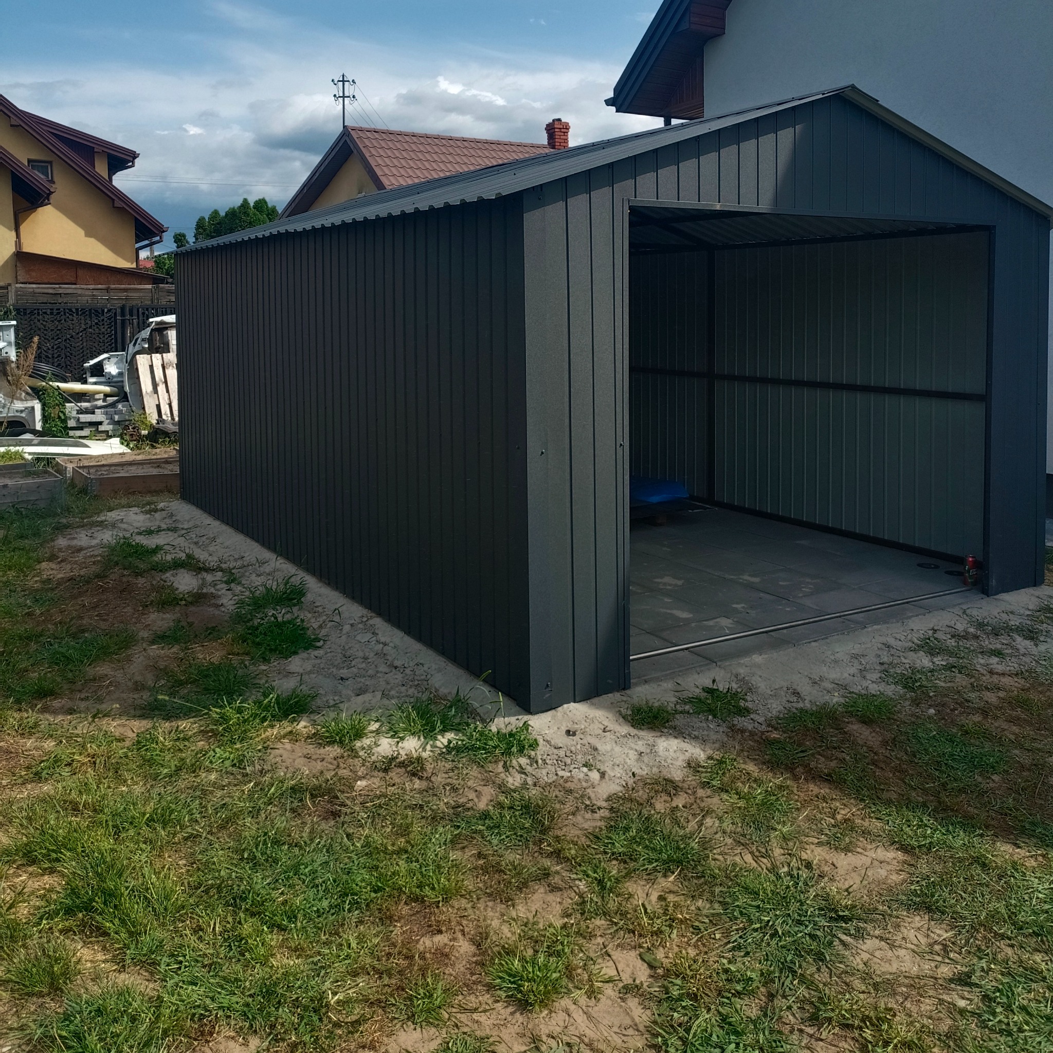 Metalowy garaż blaszany w kolorze grafitowym, z otwartymi drzwiami, na trawiastym podłożu. Widoczne budynki w tle.