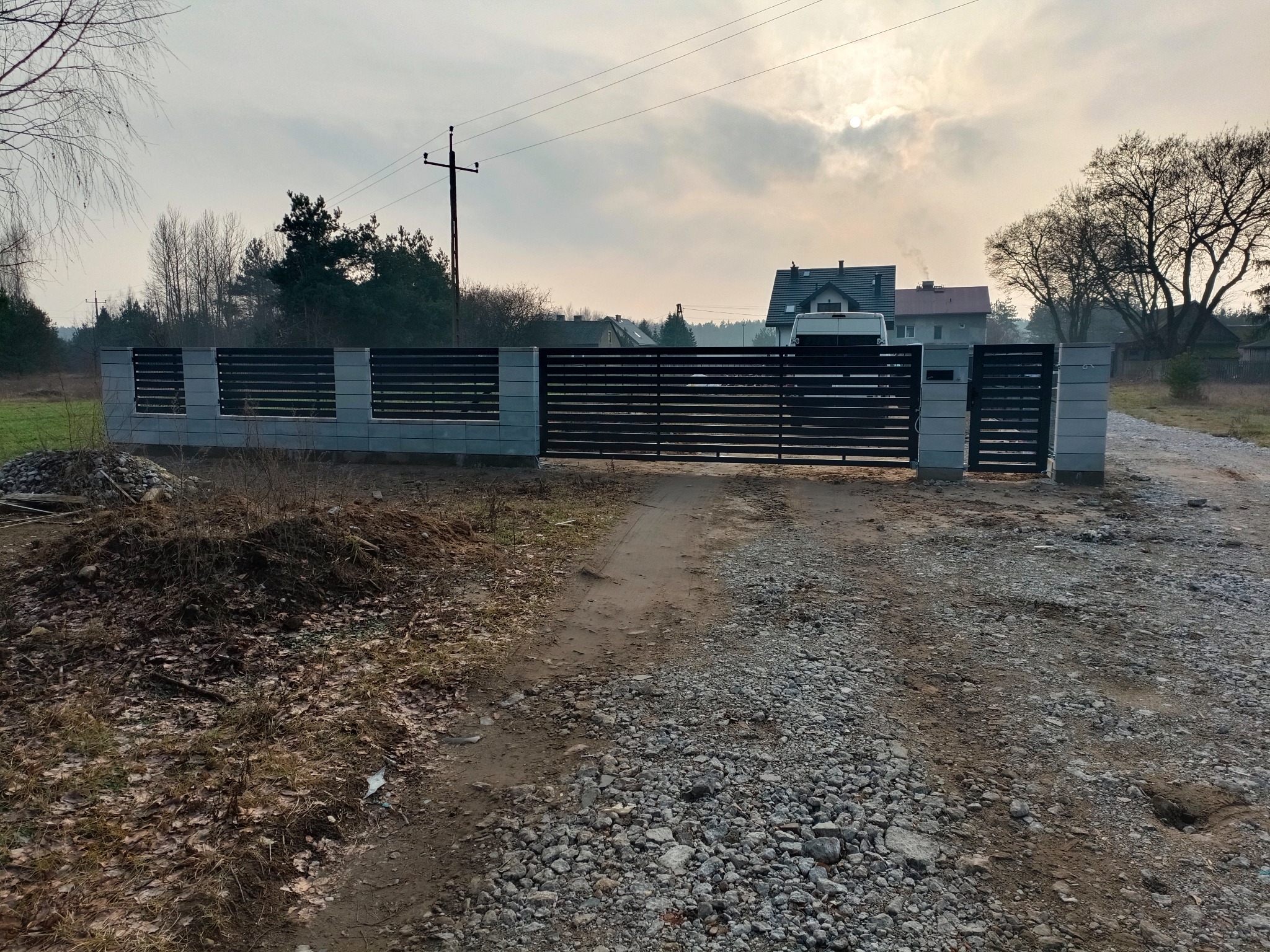 Nowoczesna brama wjazdowa z poziomymi panelami, osadzona między murowanymi słupkami. Szara kolorystyka bramy i słupków kontrastuje z naturalnym otoczeniem.