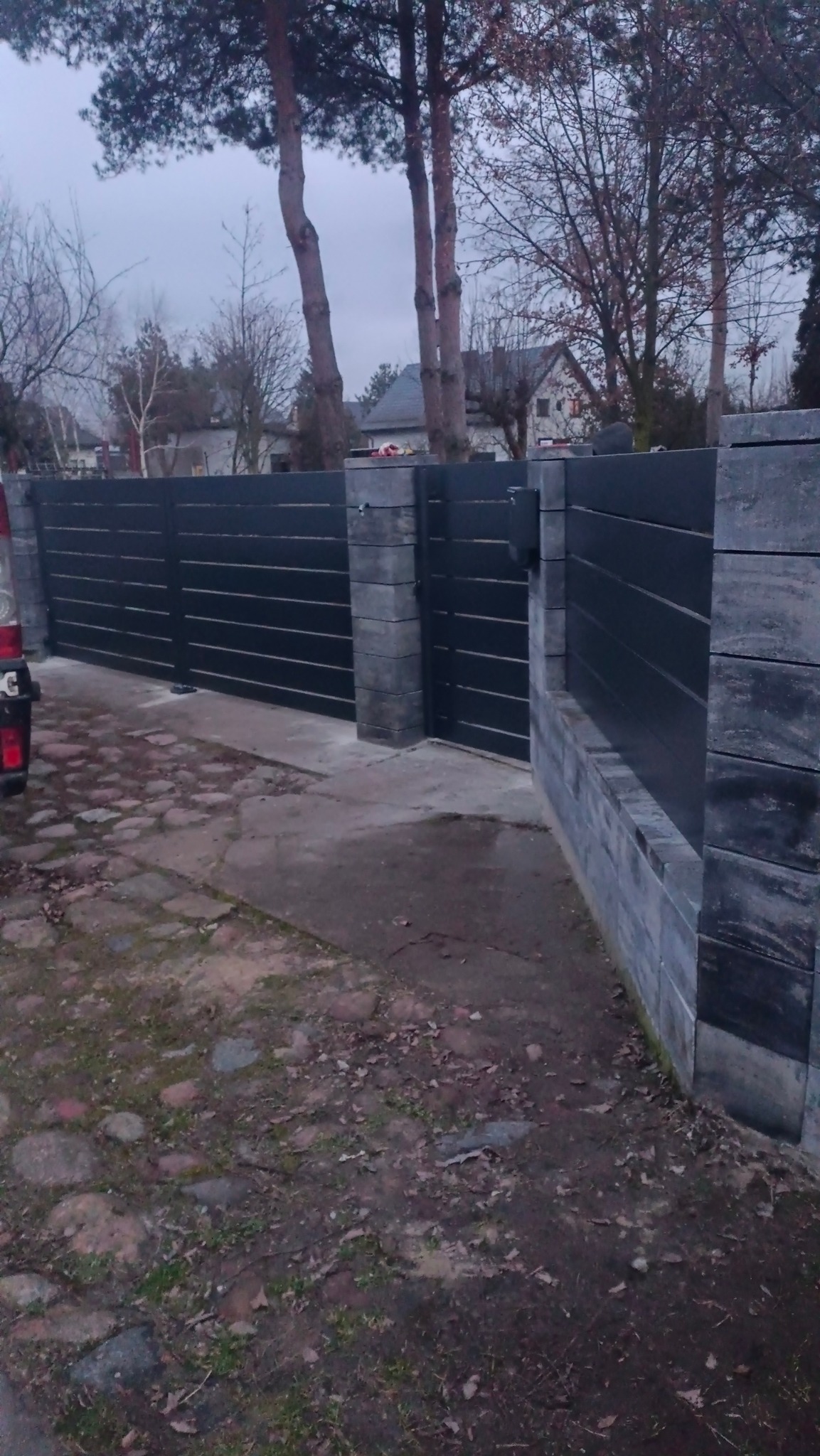 Nowoczesna, pozioma brama przesuwna w kolorze antracytowym, z elementami murowanymi z szarej kostki. Widoczny fragment chodnika z kostki brukowej i zieleń.