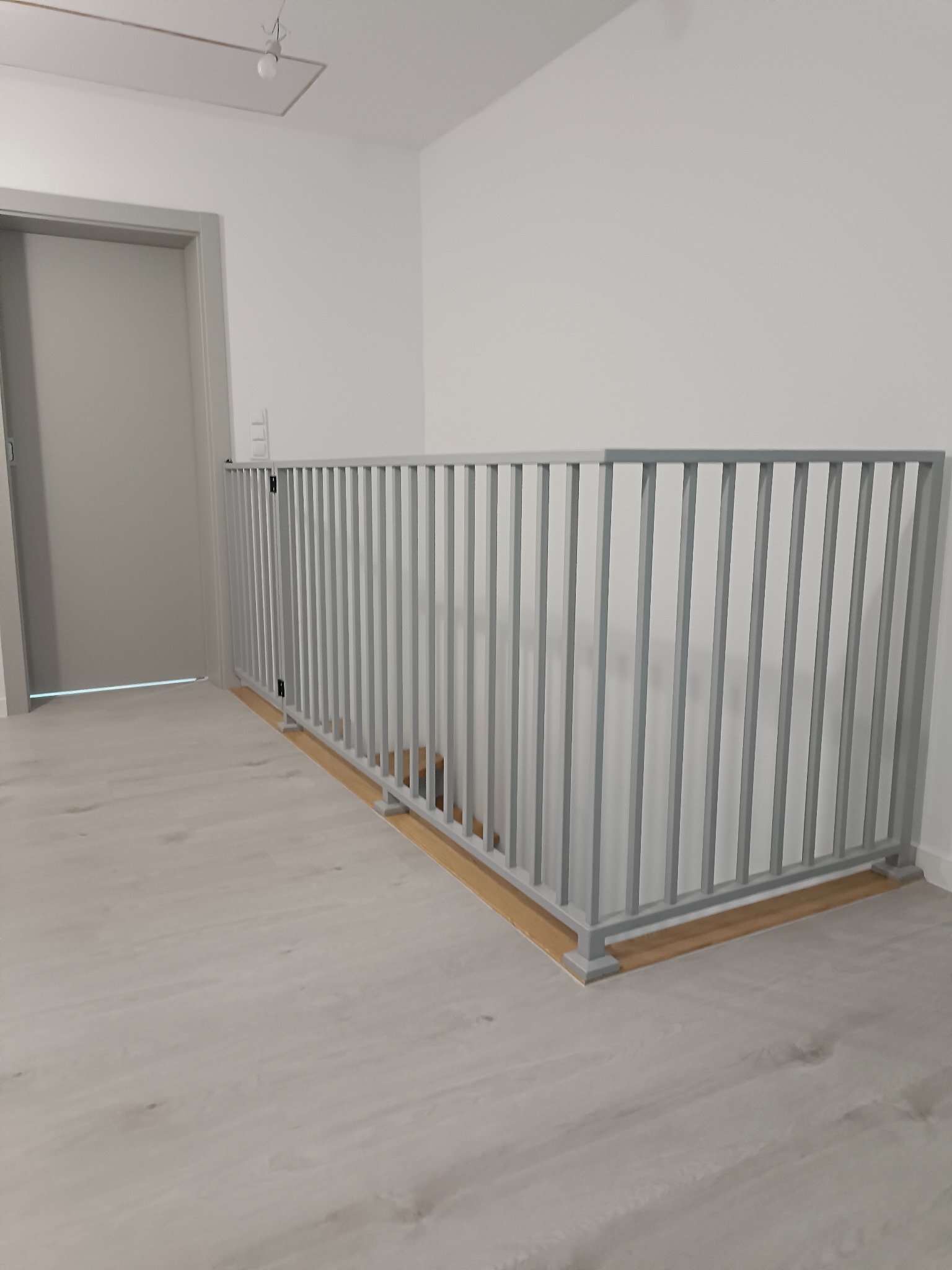 Szara, metalowa balustrada schodowa z pionowymi szczeblami, zabezpieczająca schody. Widok z boku, na jasnym tle ściany i podłogi. Minimalistyczny design.