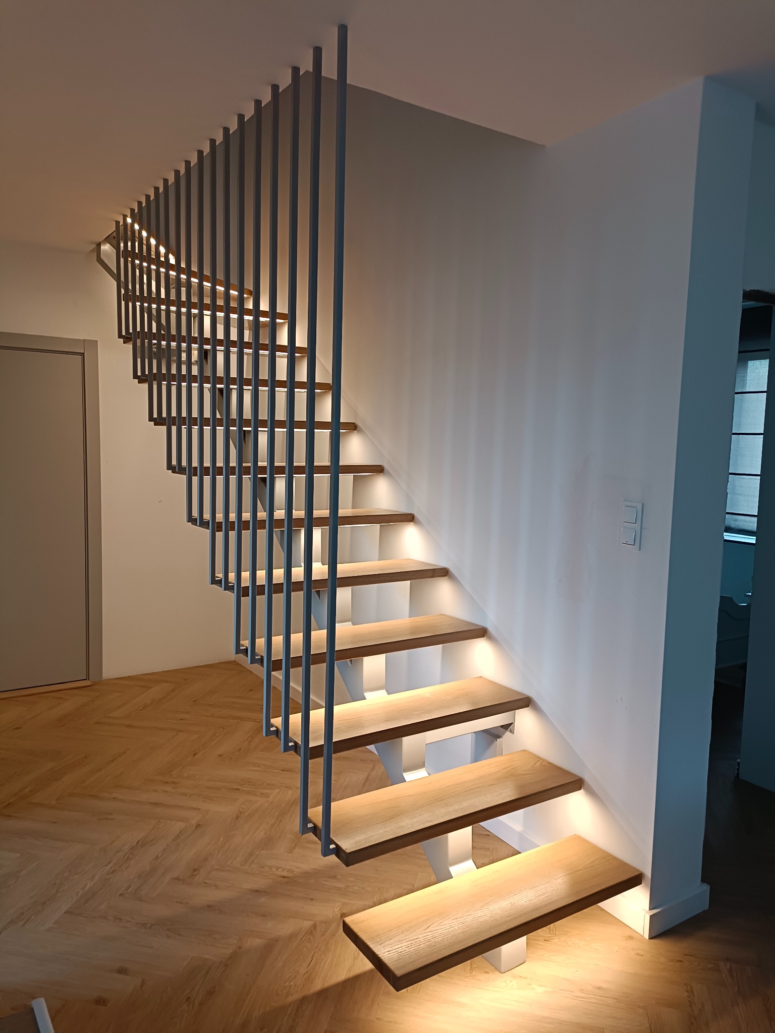 Nowoczesne schody z drewnianymi stopniami i metalową balustradą, podświetlone od spodu, na tle parkietu ułożonego w jodełkę. Minimalistyczny design.