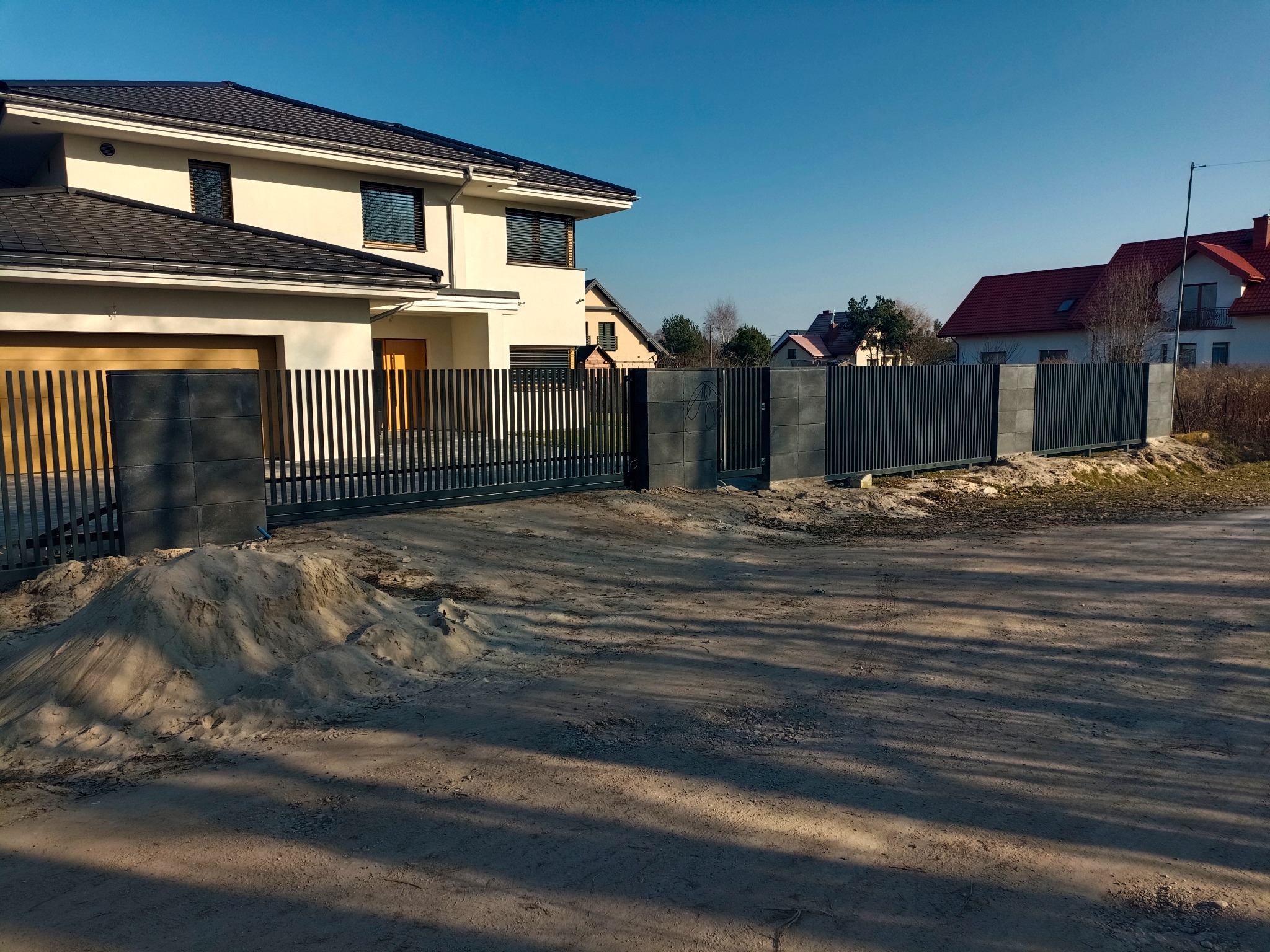 Nowoczesne ogrodzenie posesji z przesuwną bramą wjazdową i furtką, z szarymi, pionowymi panelami i betonowymi słupkami. W tle dom jednorodzinny i błękitne niebo.