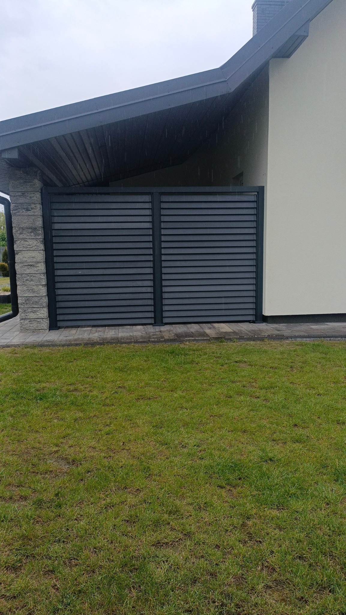 Nowoczesna, grafitowa brama lamelowa z poziomym układem desek, wkomponowana w bryłę domu z kamienną kolumną i trawnikiem. Widok z poziomu gruntu.