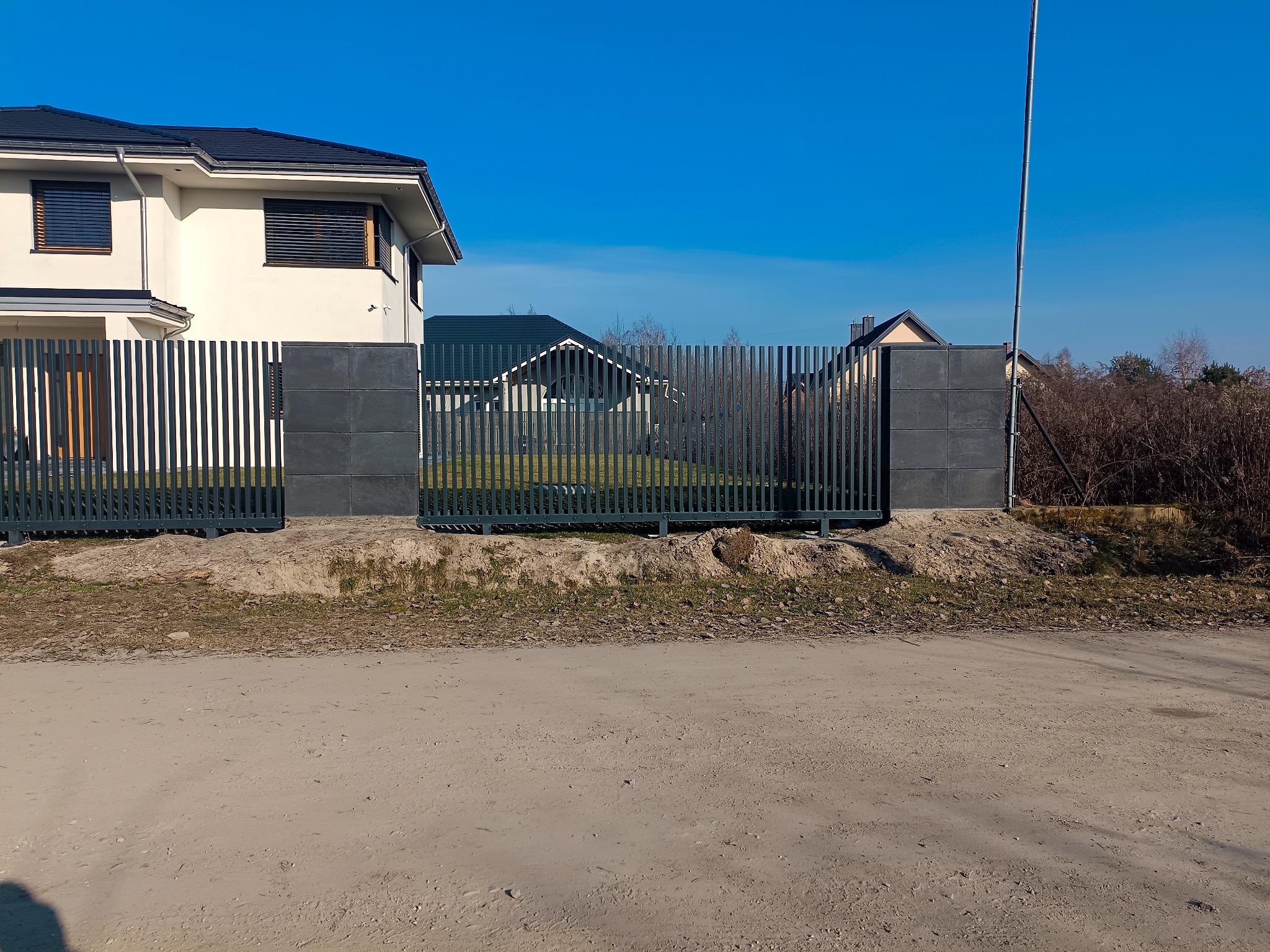 Nowoczesne ogrodzenie panelowe w kolorze grafitowym z betonowymi słupkami, na tle domu jednorodzinnego i błękitnego nieba. Widok z zewnątrz posesji.