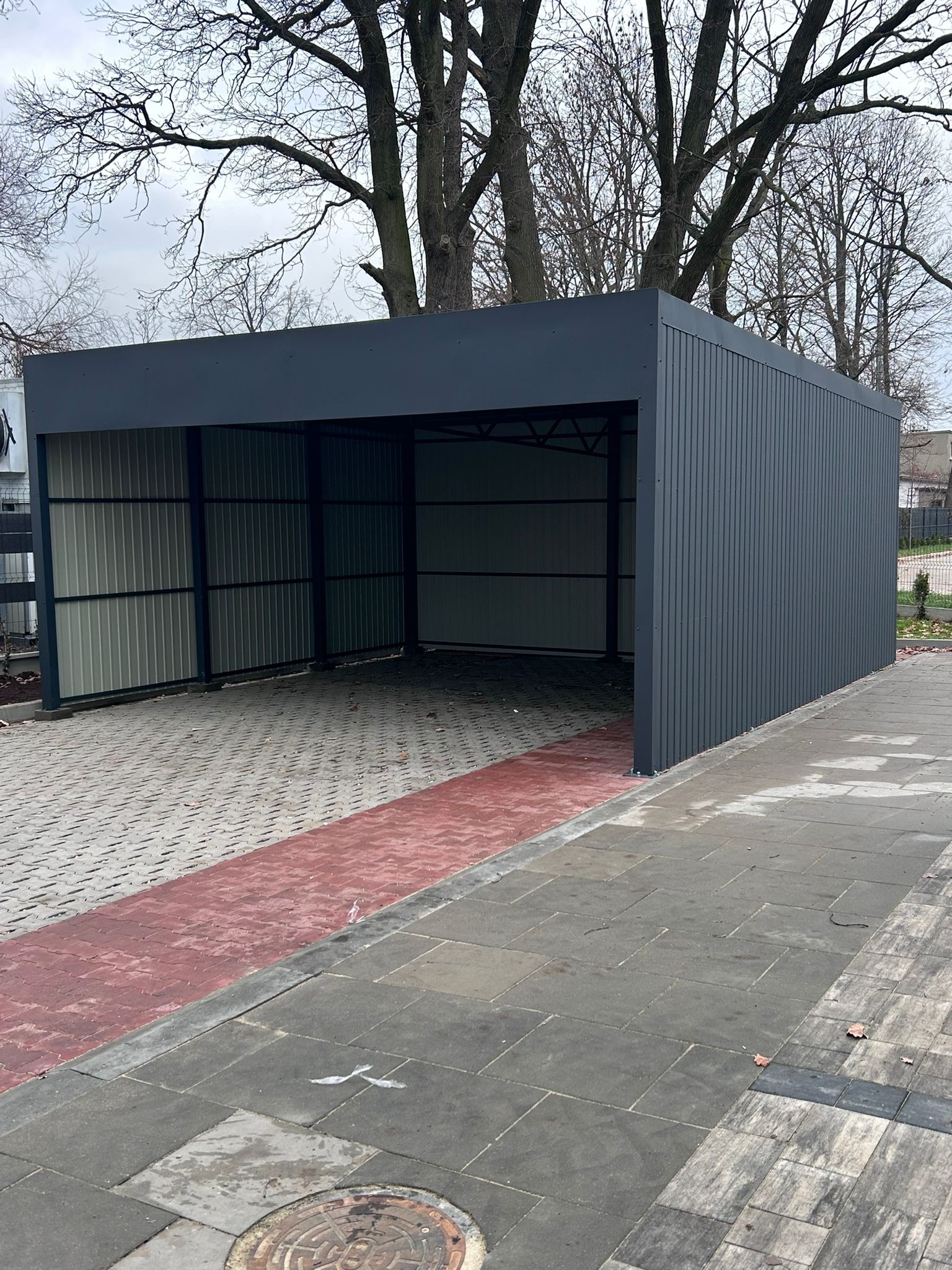 Nowoczesna, dwustanowiskowa wiata garażowa z szarej blachy trapezowej, na kostce brukowej. Widoczne drzewa w tle. Minimalistyczny design, proste linie.