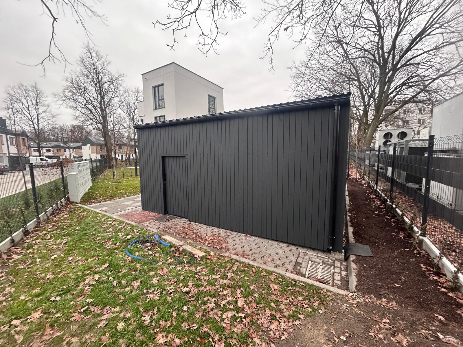 Ciemnoszara, pionowo panelowa konstrukcja z drzwiami, obok czarny panelowy płot i trawnik z liśćmi. W tle budynek mieszkalny i drzewa bez liści.