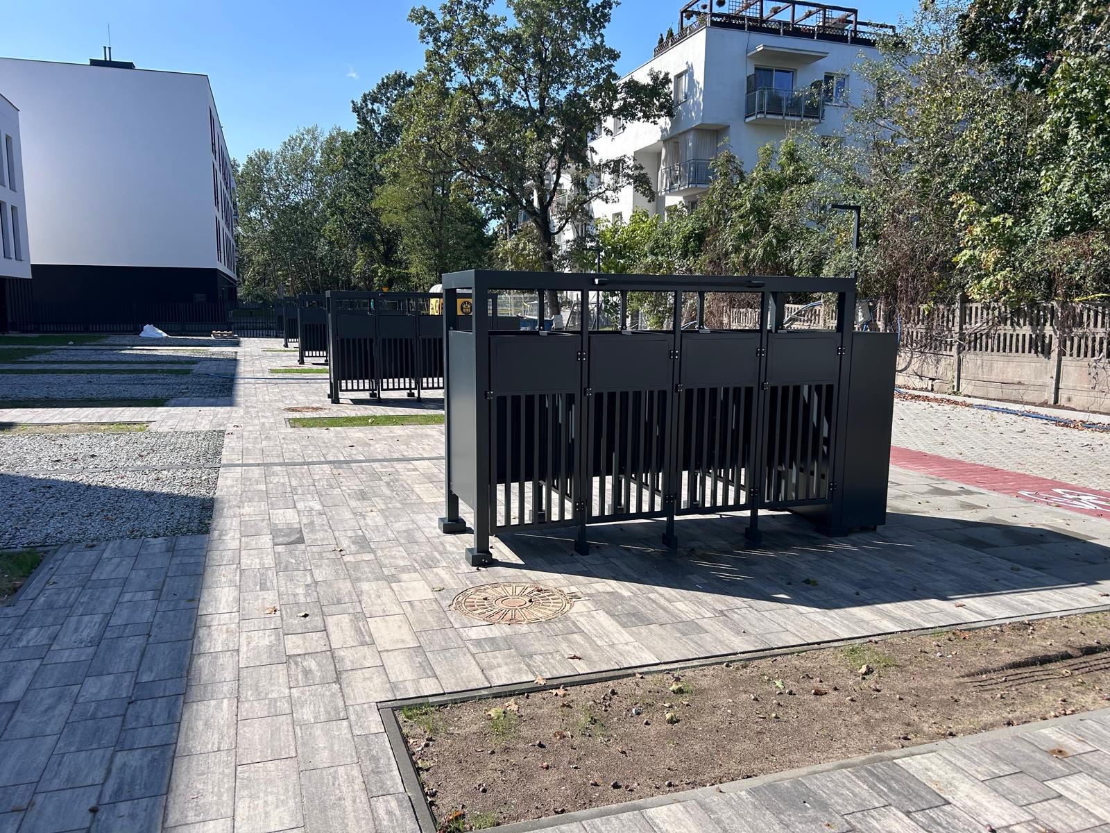 Szereg czarnych, metalowych barierek z pionowymi szczebelkami, ustawionych wzdłuż ścieżki z kostki brukowej na tle nowoczesnych budynków i zieleni. Słoneczny dzień.