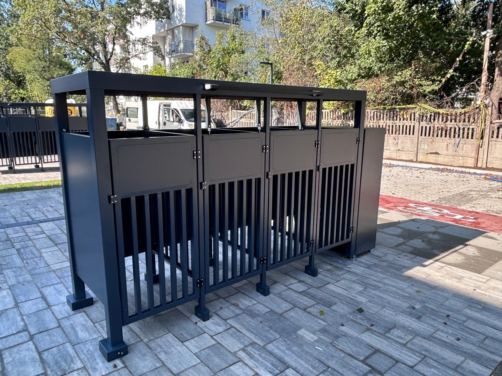 Ciemnoszara, metalowa konstrukcja z pionowymi szczebelkami, prawdopodobnie osłona na śmietniki, na tle chodnika z kostki i zieleni miejskiej.