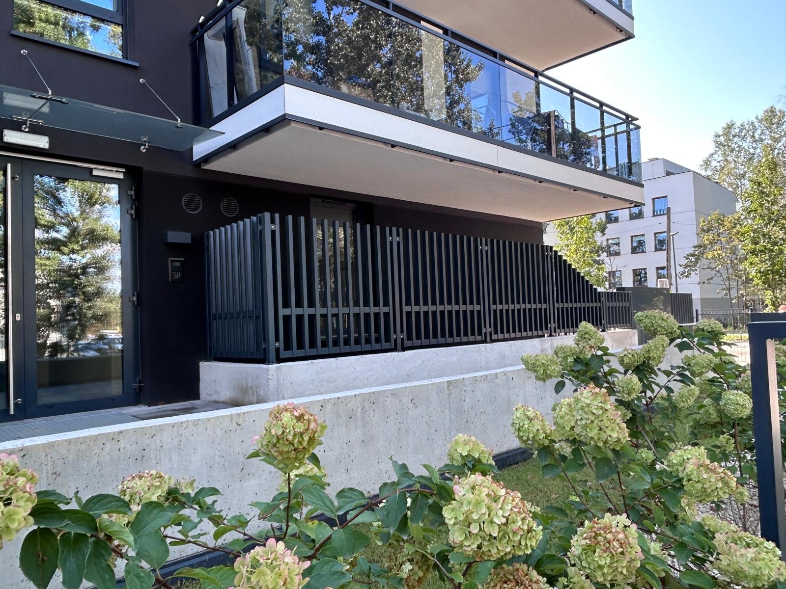 Nowoczesna, czarna balustrada balkonowa z pionowych profili, zamontowana na betonowej podmurówce. W tle budynek z przeszklonymi balustradami i zielenią.