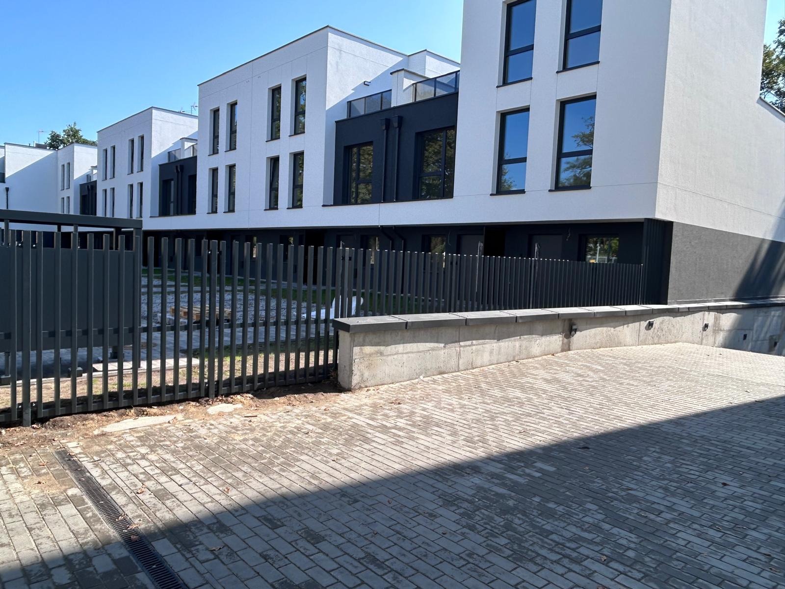 Nowoczesne, grafitowe ogrodzenie panelowe przed szeregiem budynków w jasnej kolorystyce. Widoczny fragment chodnika z kostki brukowej i betonowy murek.