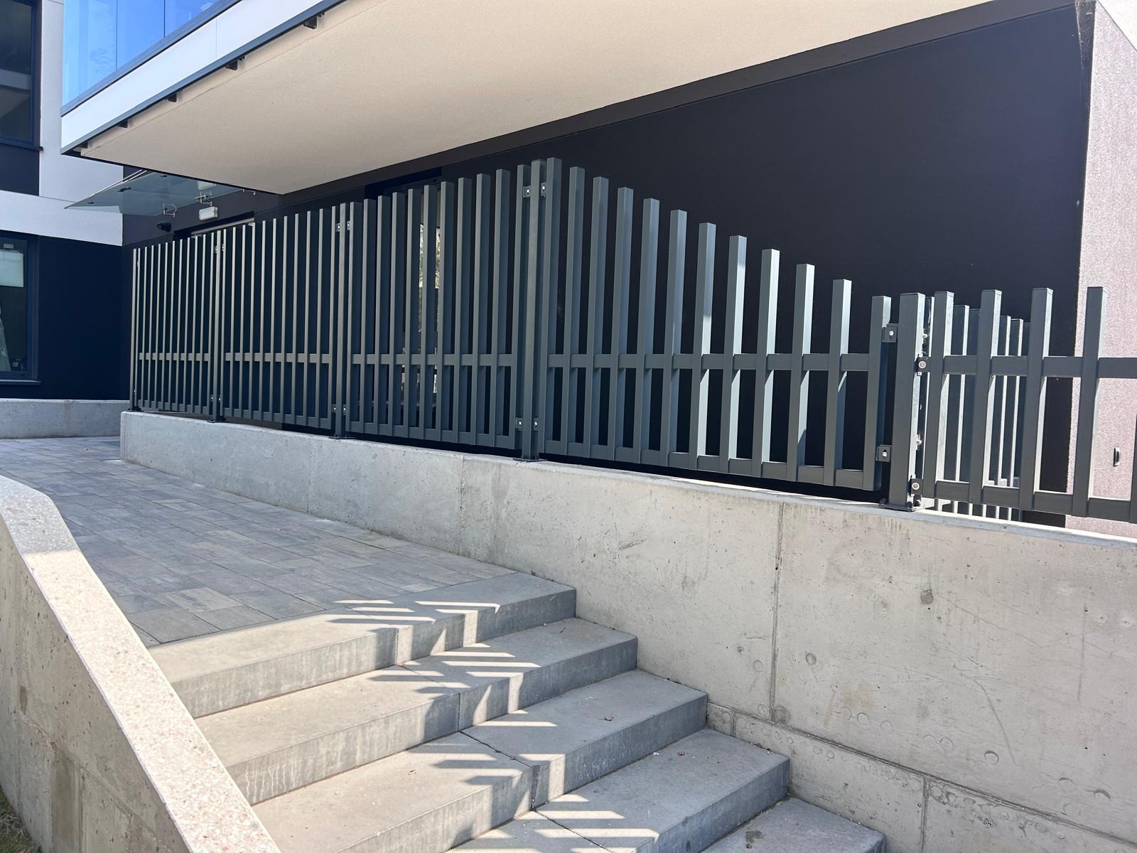 Nowoczesne, metalowe ogrodzenie w kolorze grafitowym, zamontowane na betonowym murku przy schodach wejściowych do budynku. Minimalistyczny design z pionowymi elementami.