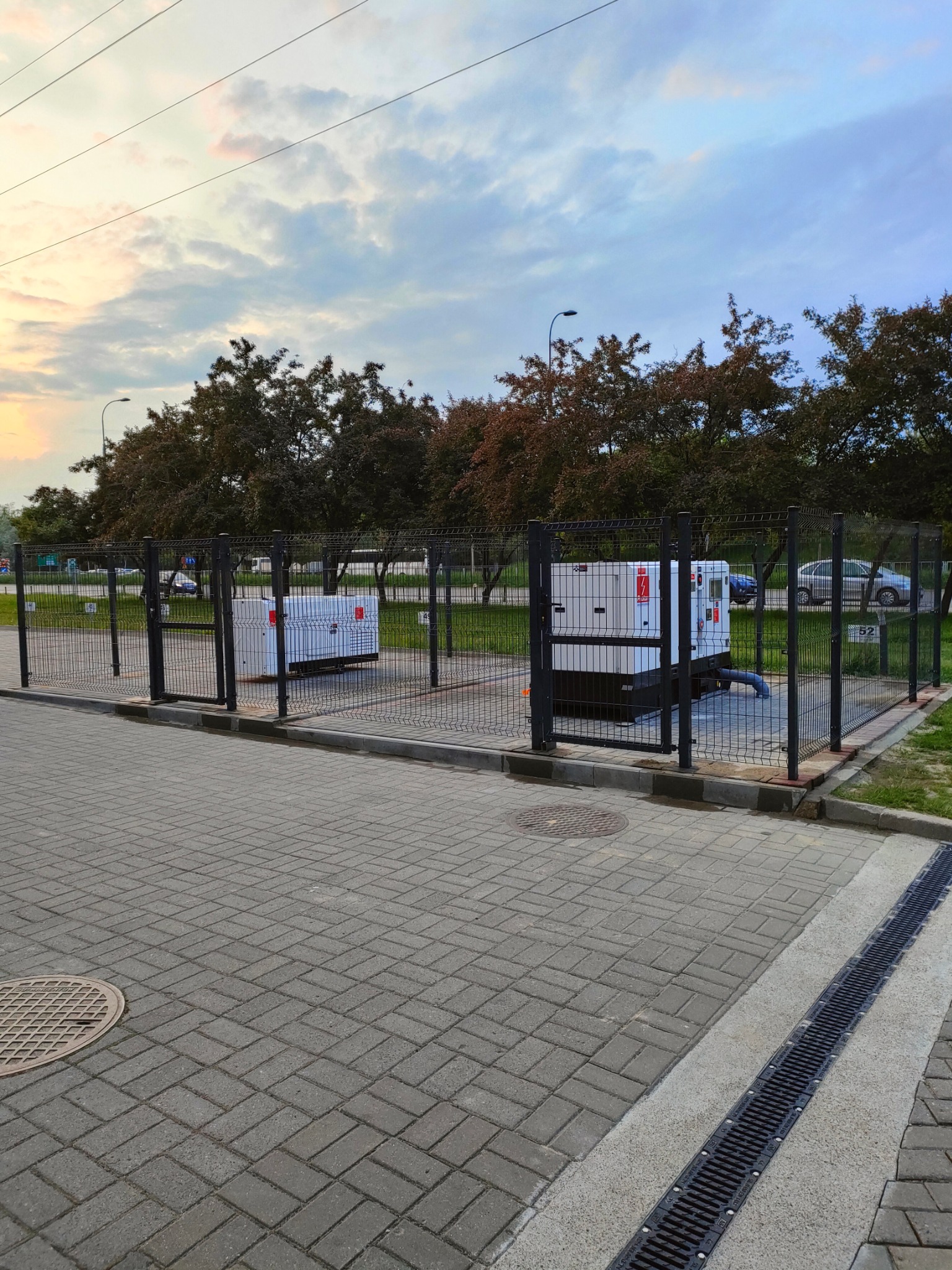 Dwa agregaty prądotwórcze umieszczone za czarnym, metalowym ogrodzeniem panelowym na zewnątrz budynku; widoczne drzewa i zachmurzone niebo w tle.