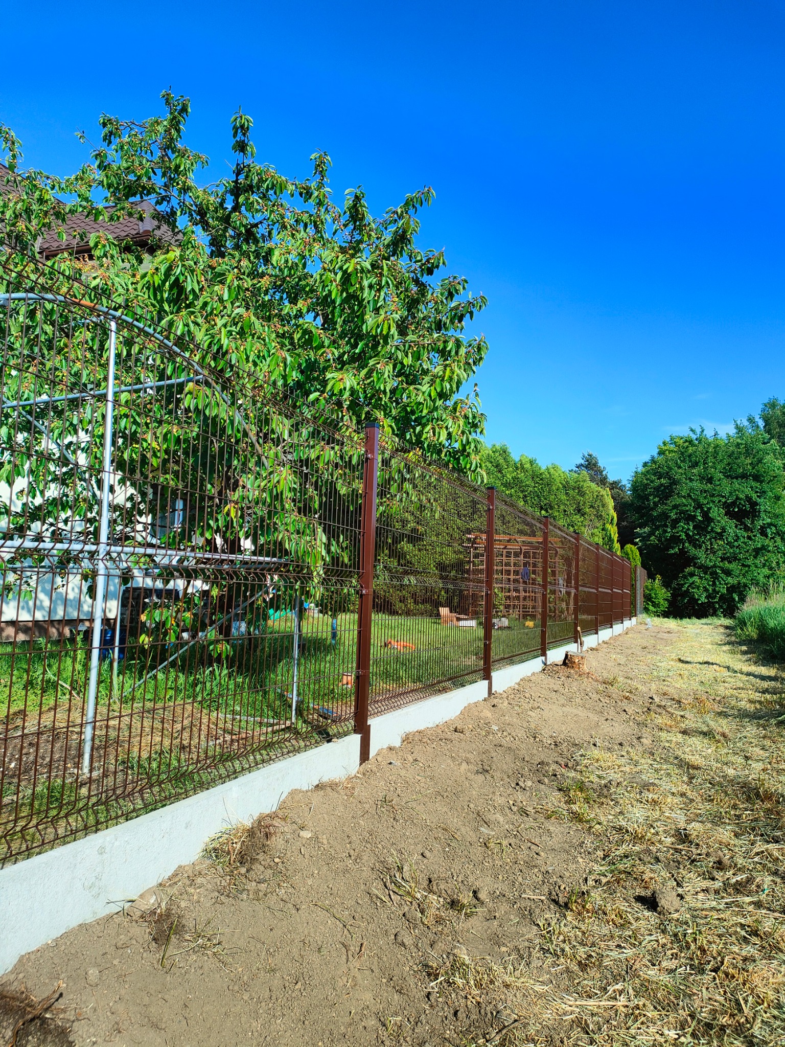 Nowo zainstalowane brązowe ogrodzenie panelowe z podmurówką betonową, widok z zewnątrz posesji, z drzewami w tle i błękitnym niebem.