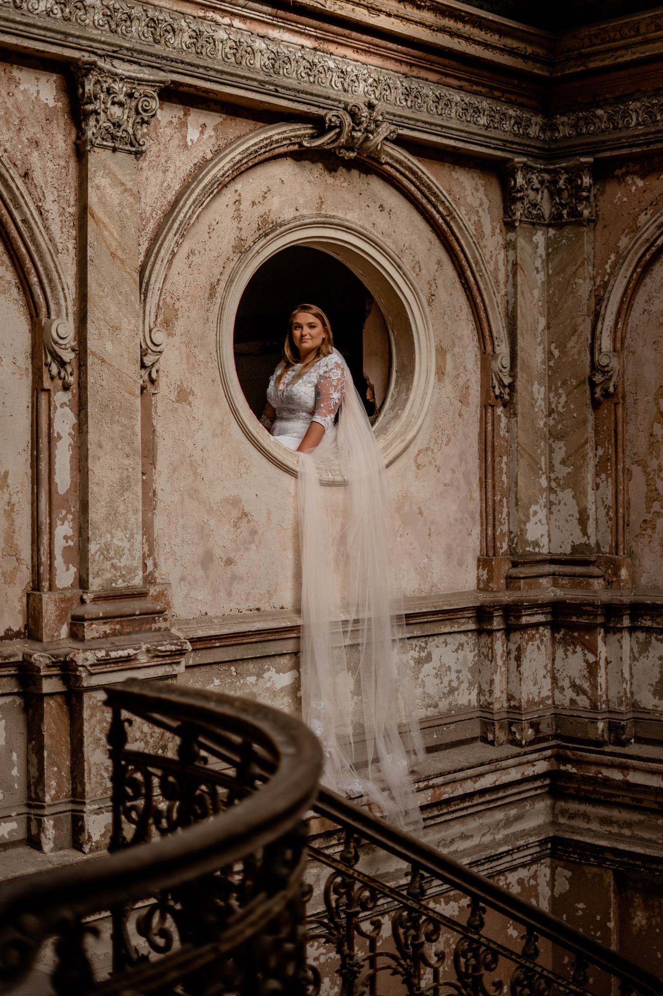 Kobieta w koronkowej sukni ślubnej i welonie pozuje w okrągłym oknie starego, zaniedbanego budynku z ozdobnymi detalami architektonicznymi i schodami z kutą balustradą.