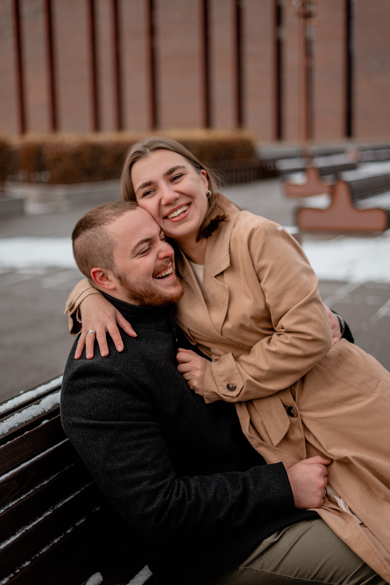 Uśmiechnięta para narzeczonych w jesiennych strojach, obejmująca się na ławce z resztkami śniegu, w tle budynek z cegły.