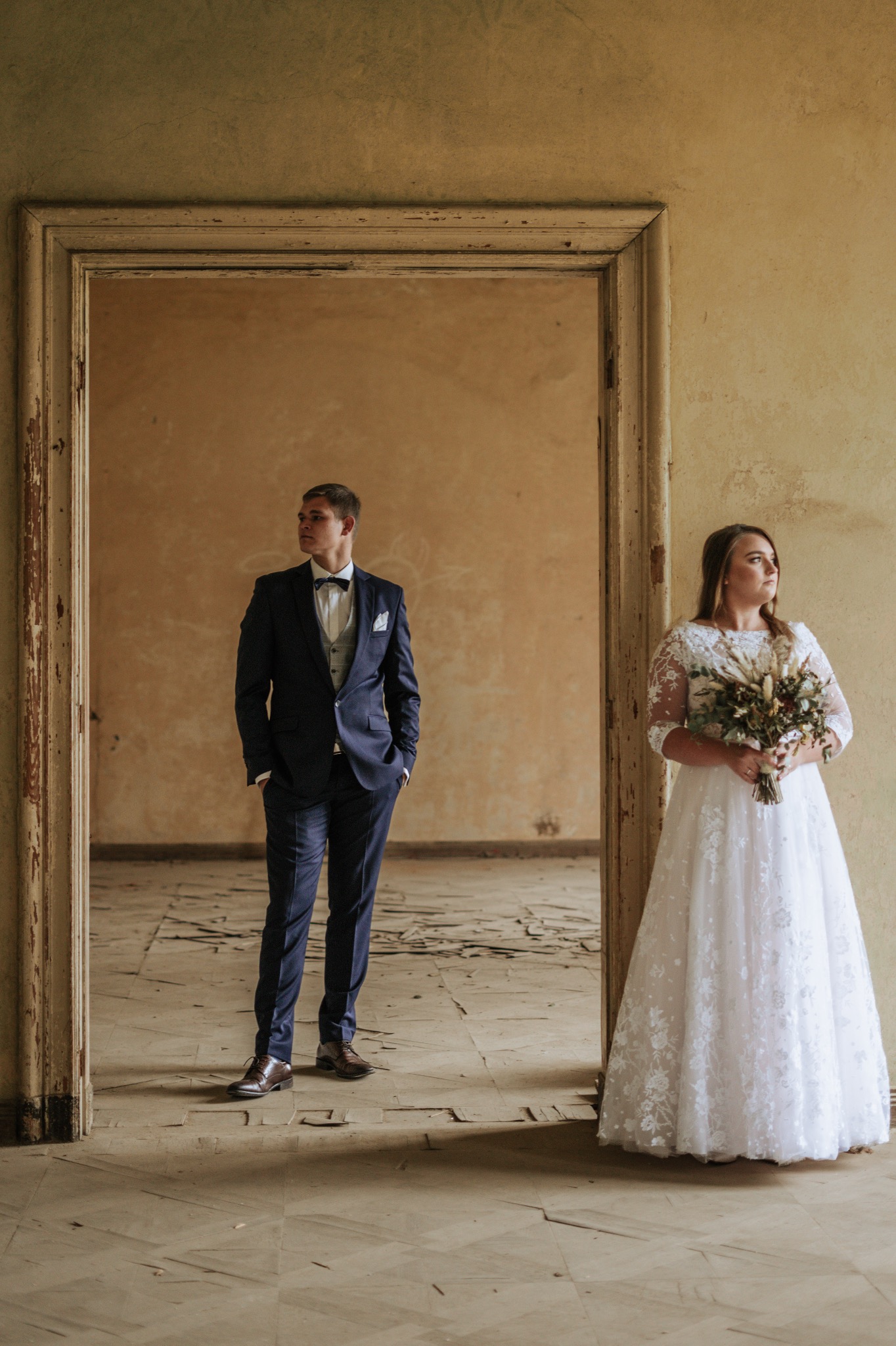 Para młoda w opuszczonym budynku: panna młoda w koronkowej sukni trzyma bukiet suszonych kwiatów, pan młody w granatowym garniturze stoi w przejściu.