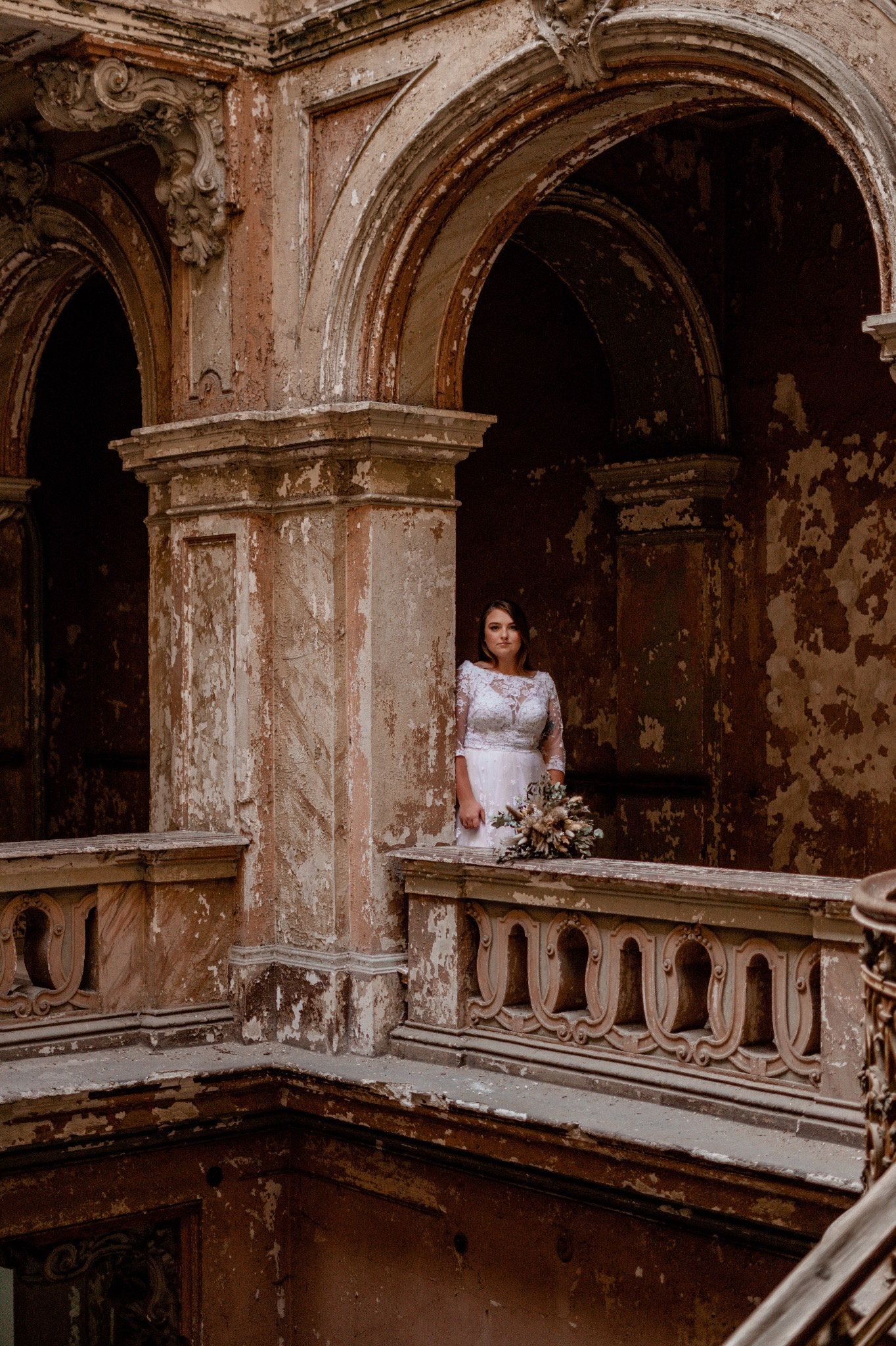Panna młoda w koronkowej sukni z bukietem na balkonie w opuszczonym pałacu z łuszczącą się farbą i marmurowymi kolumnami.