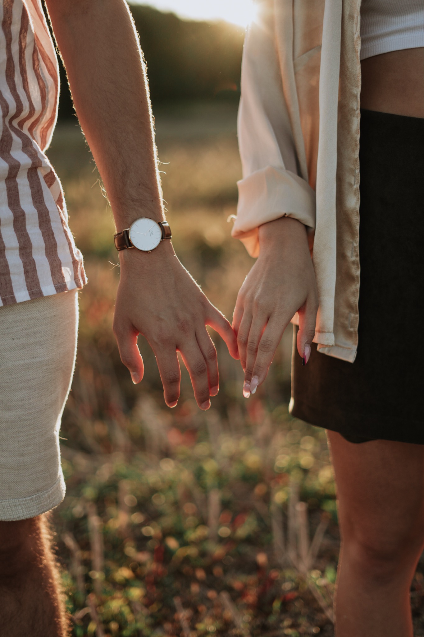 Para trzymająca się za ręce w promieniach zachodzącego słońca, widoczny zegarek na nadgarstku mężczyzny i manicure u kobiety.