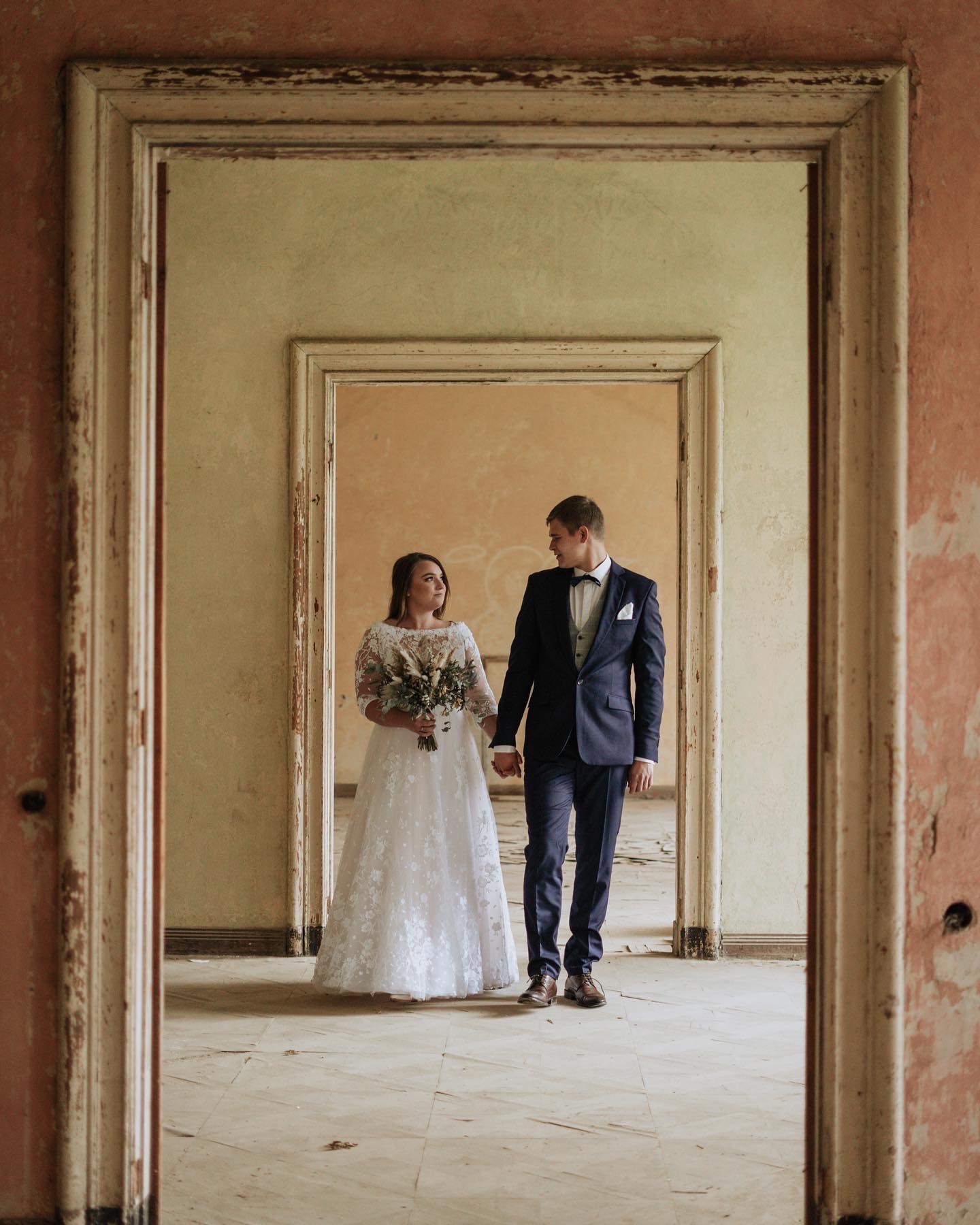 Para młoda trzymająca się za ręce, idąca przez serię zabytkowych, zniszczonych futryn drzwiowych, panna młoda w koronkowej sukni, pan młody w granatowym garniturze z muchą.