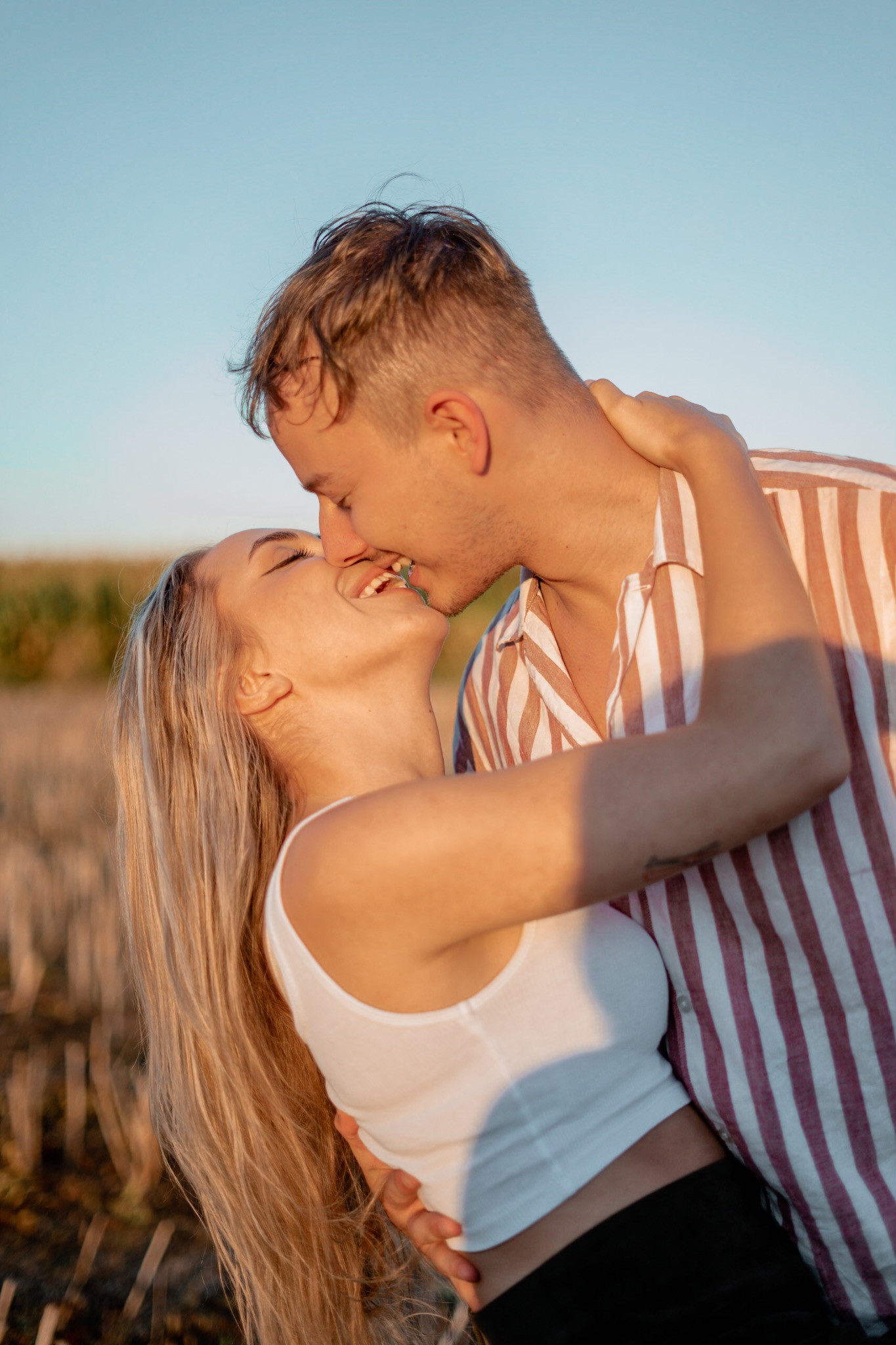 Zakochana para w objęciach, pocałunek na tle pola w ciepłym świetle zachodzącego słońca, ona w białym topie, on w koszuli w paski.