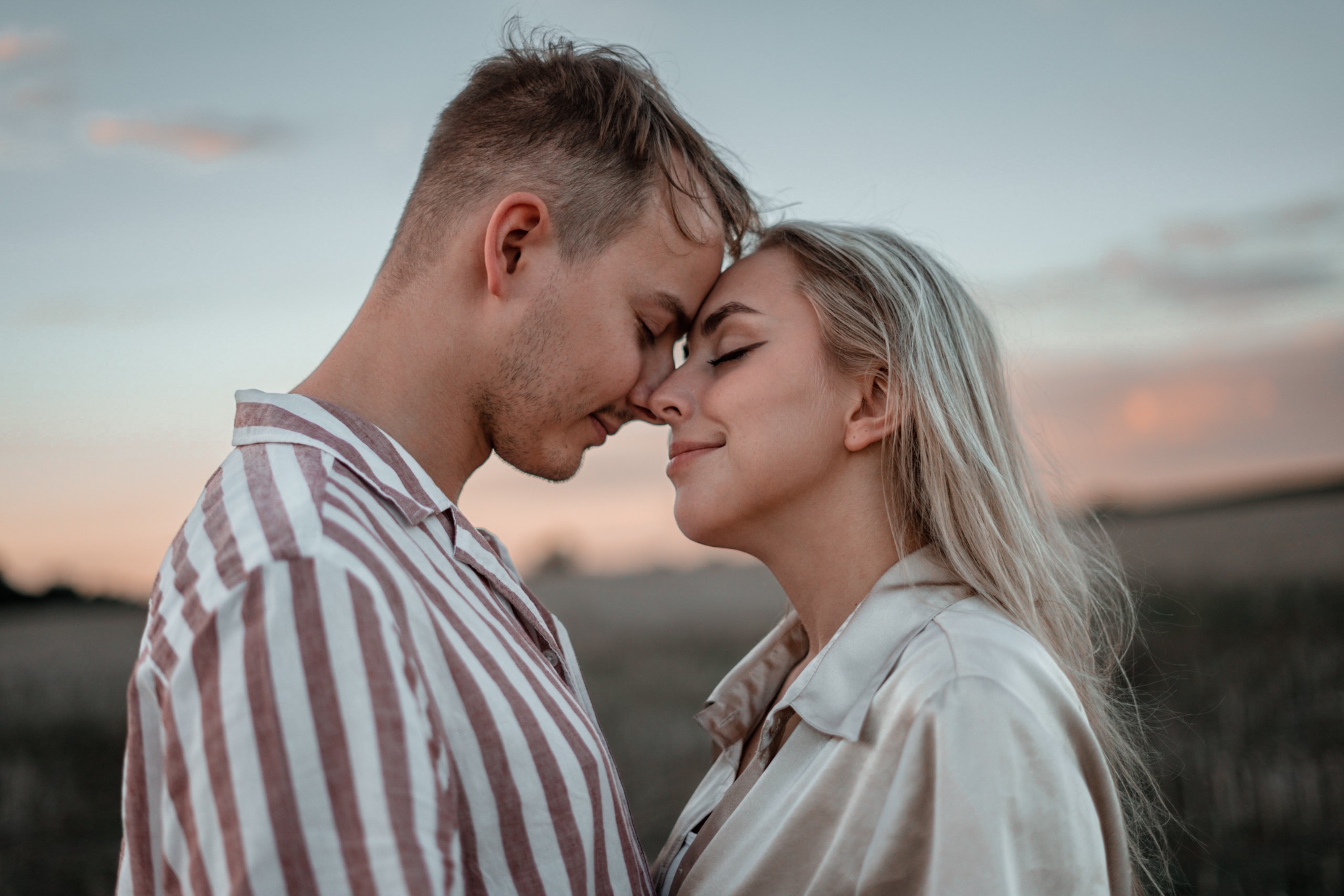 Młoda para, mężczyzna w koszuli w paski i kobieta z blond włosami w satynowej bluzce, dotykają się czołami na tle zachodzącego słońca.