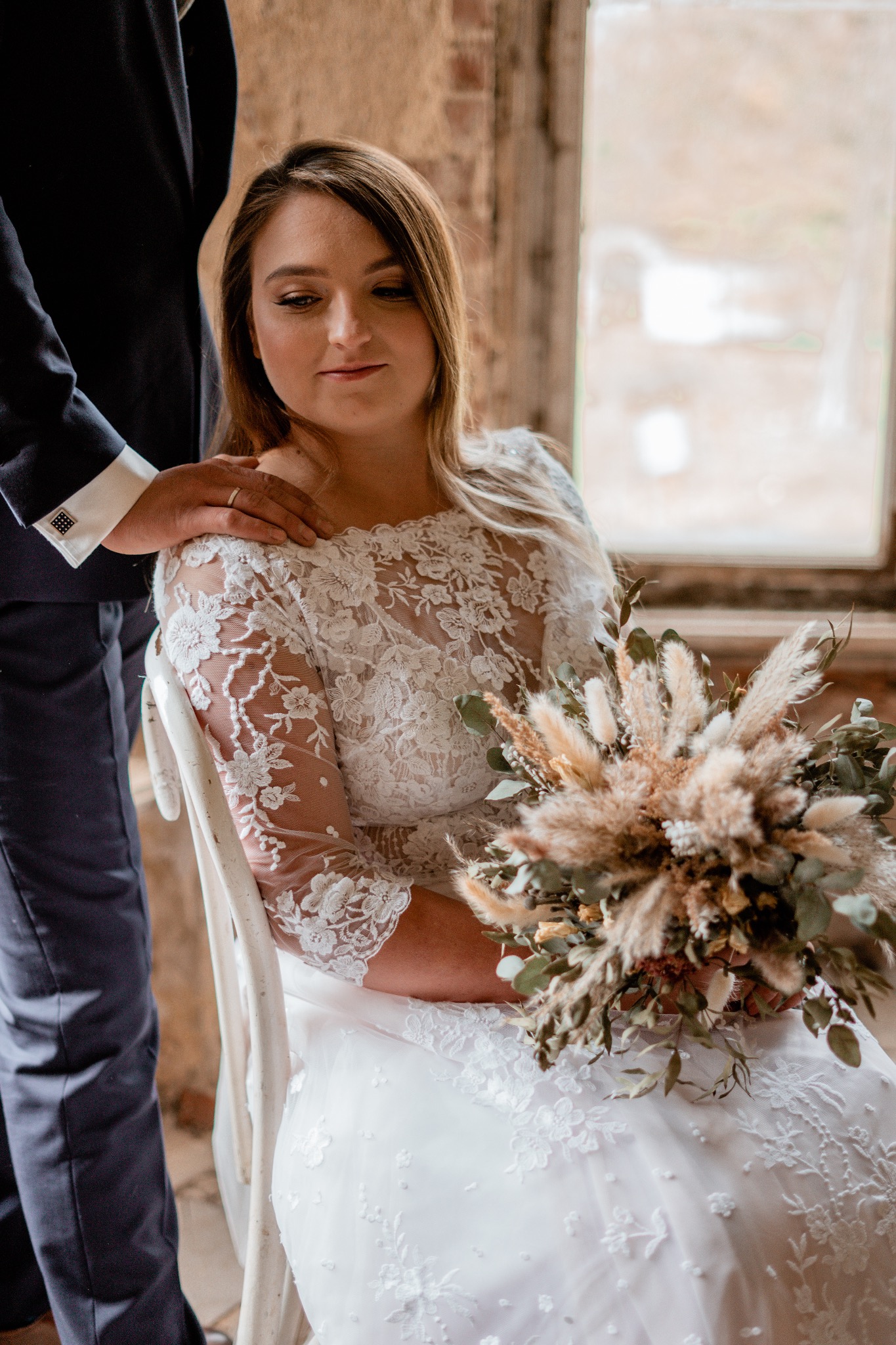 Panna młoda w koronkowej sukni siedzi na krześle, trzyma bukiet z suszonych kwiatów, obok stoi mężczyzna w garniturze z ręką na jej ramieniu.