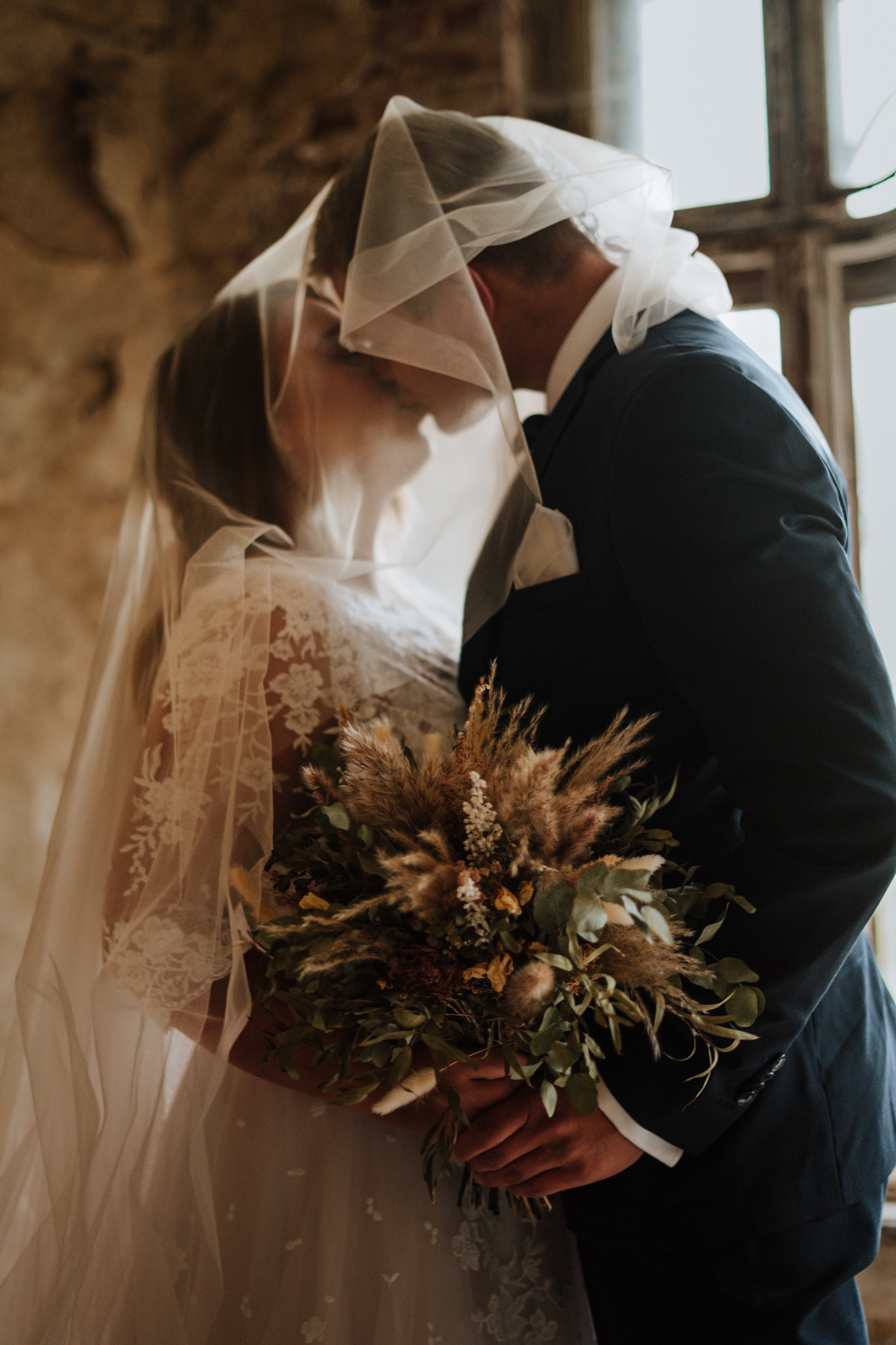 Pocałunek młodej pary pod welonem na tle okna, panna młoda trzyma bukiet z suszonych kwiatów i zieleni.