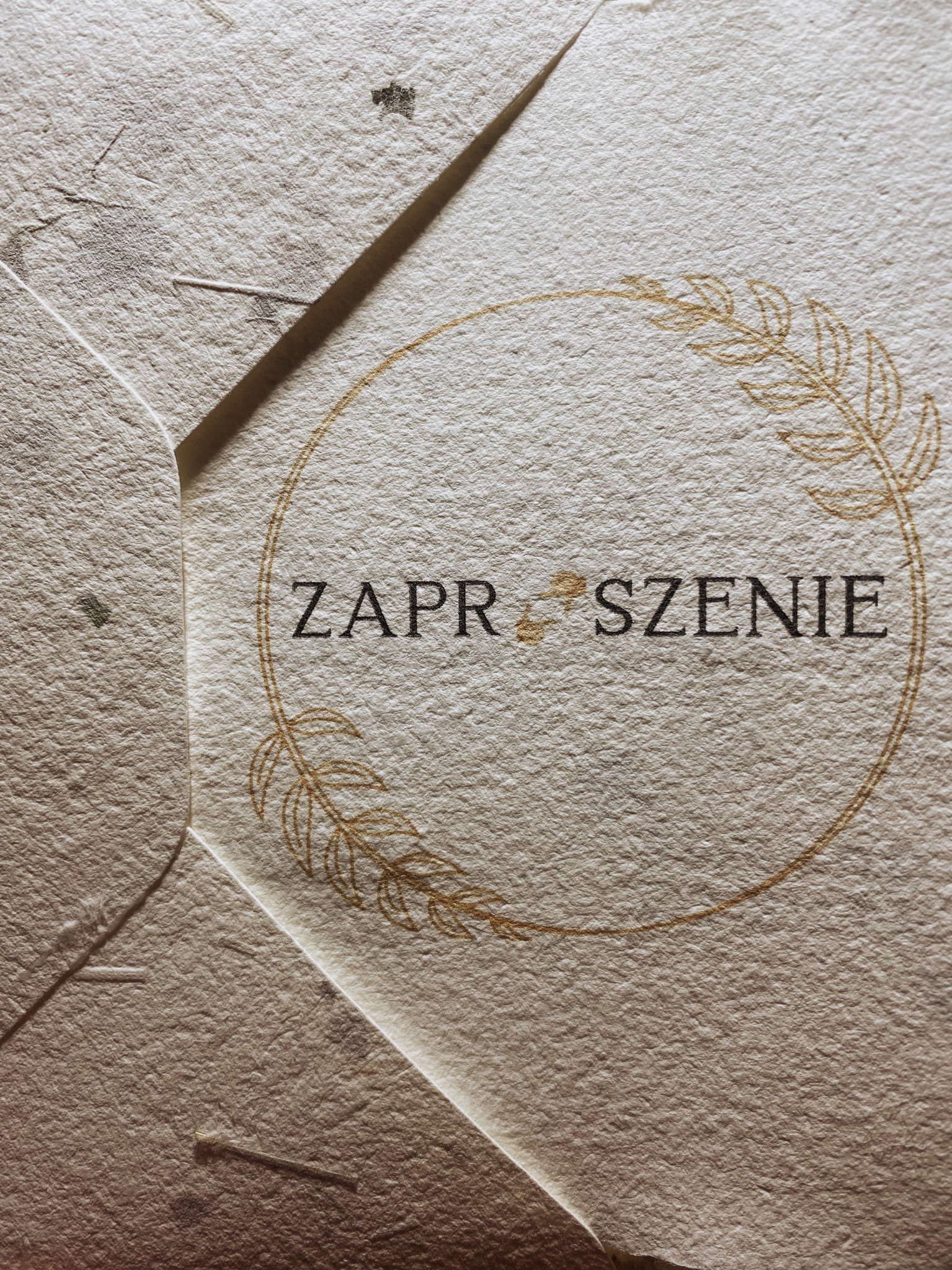 Kremowa koperta z papieru czerpanego z napisem 'Zaproszenie' otoczonym złotym wieńcem z liści