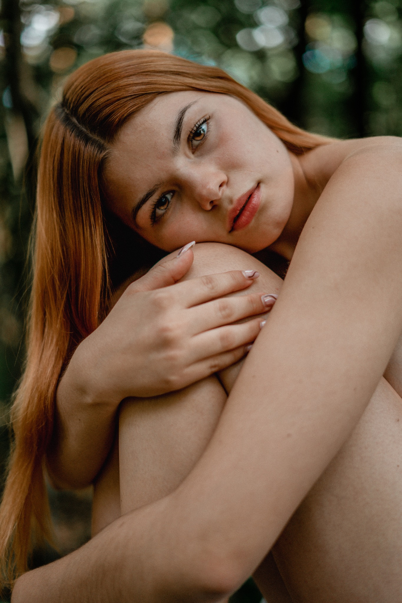 Portret kobiety z rudymi włosami, obejmującej kolana na tle zieleni, z delikatnym makijażem i jasnymi paznokciami.