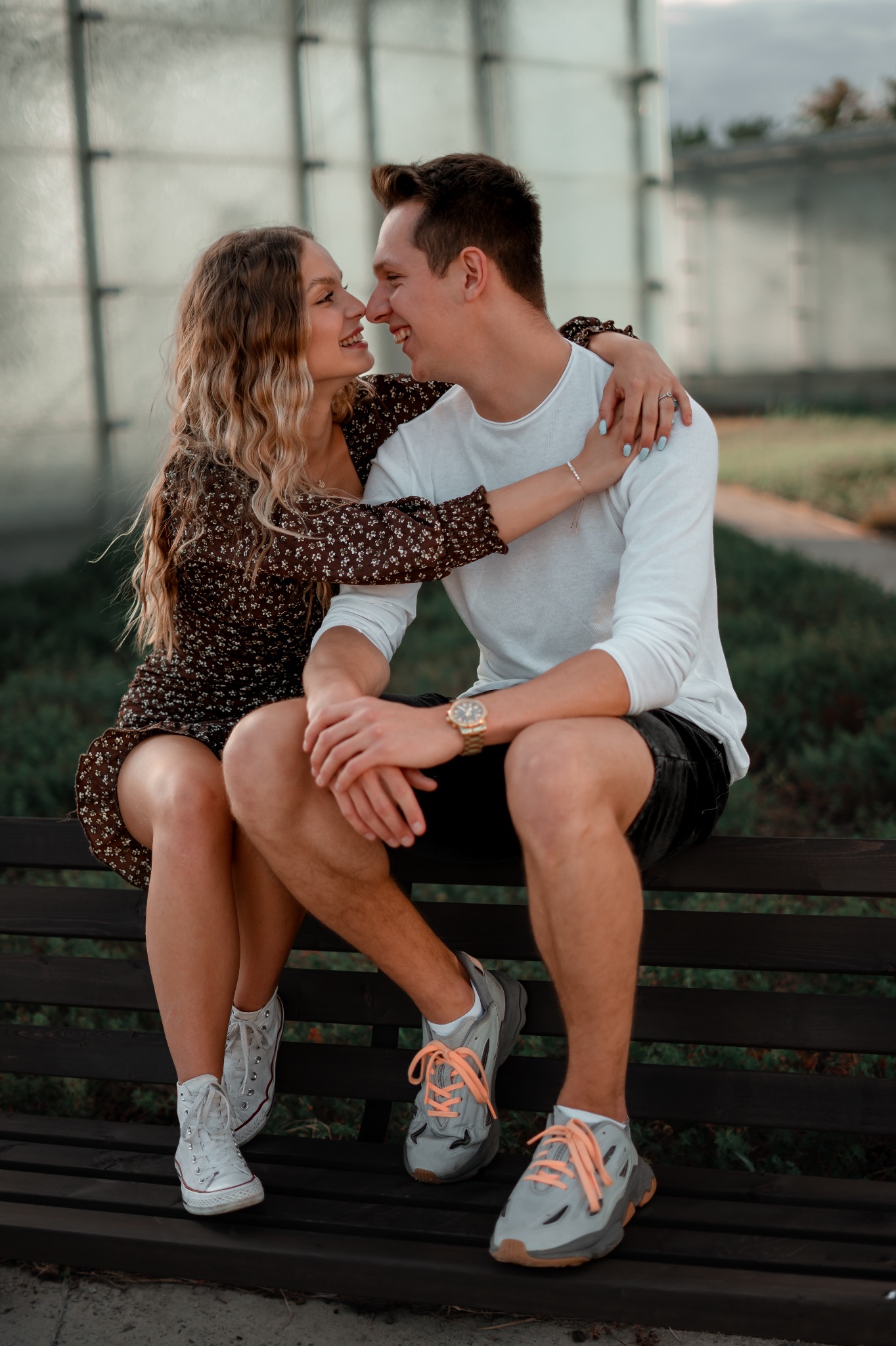 Uśmiechnięta para siedzi na ławce, dotykając się nosami, ona w kwiecistej sukience i białych trampkach, on w białej koszulce, czarnych spodenkach i szarych butach sportowych z pomarańczowymi...