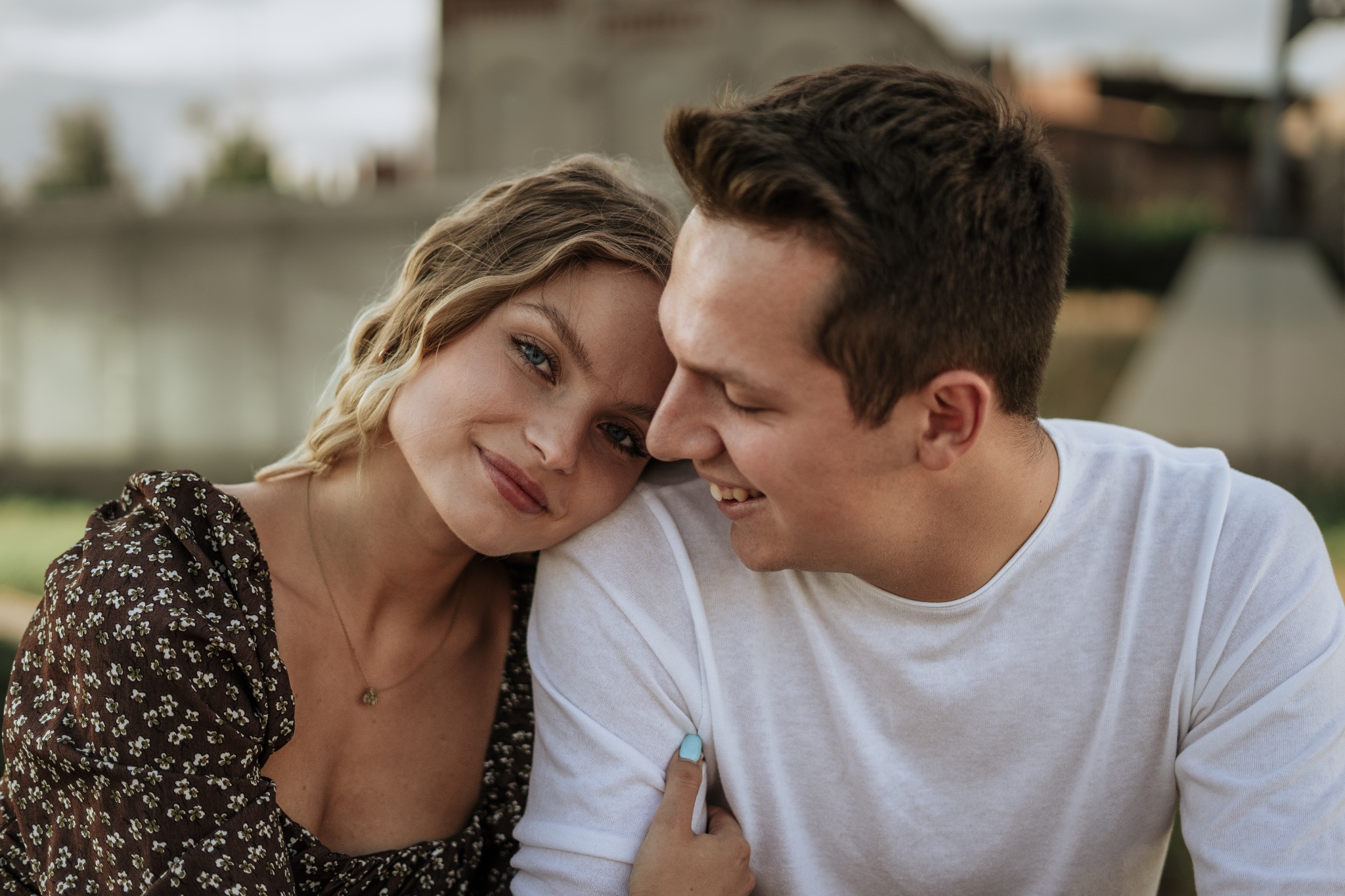 Uśmiechnięta para, ona z blond lokami w kwiecistej sukience, opiera głowę o jego ramię w białej koszulce, delikatny makijaż podkreśla jej niebieskie oczy.
