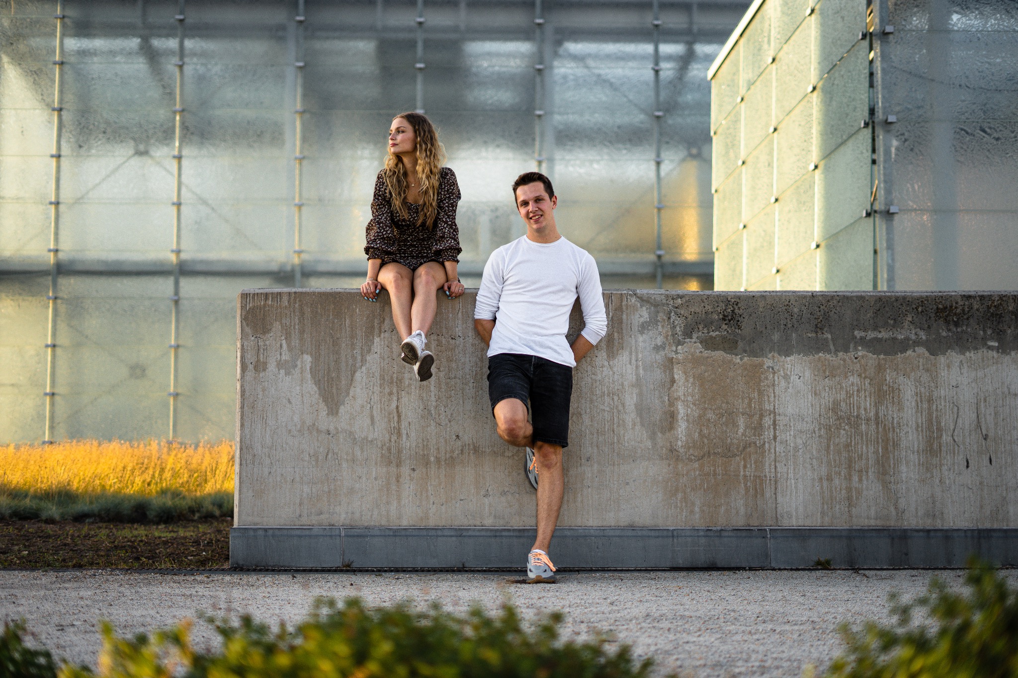 Para: blondynka siedząca na betonowym murku, chłopak oparty o mur, na tle szklanego budynku w ciepłym świetle.