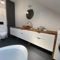 Nowoczesna łazienka na poddaszu z białą umywalką w kolorze bursztynu, długą szafką z białymi frontami i drewnianym blatem, białą pralką Samsung i owalnym lustrem.