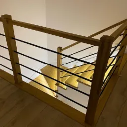 Drewniana balustrada z czarnymi, poziomymi prętami, oświetlone schody w tle, widok z góry na drewnianą podłogę.