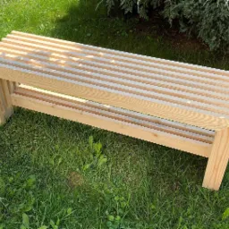 Surowa, drewniana ławka ogrodowa z listew, ustawiona na trawie w słoneczny dzień, z krzewem w tle.