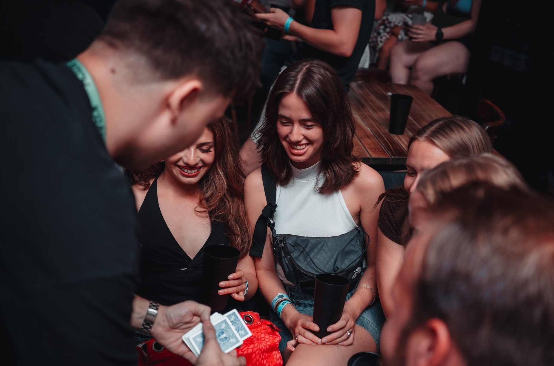 Uśmiechnięte kobiety obserwują trik karciany wykonywany przez iluzjonistę w ciemnym pomieszczeniu. Widoczne karty, napoje w czarnych kubkach i fragmenty innych osób.