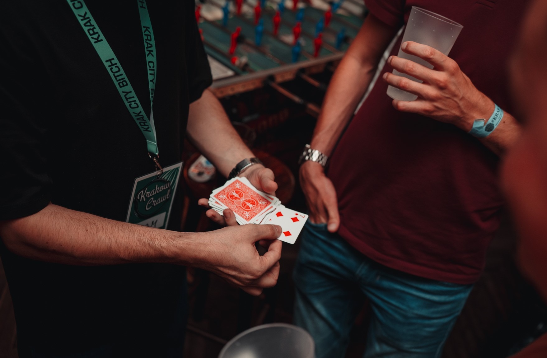 Karty w dłoniach iluzjonisty podczas występu, w tle stół do gry i osoba z napojem. Widoczny identyfikator Krakow Crawl.