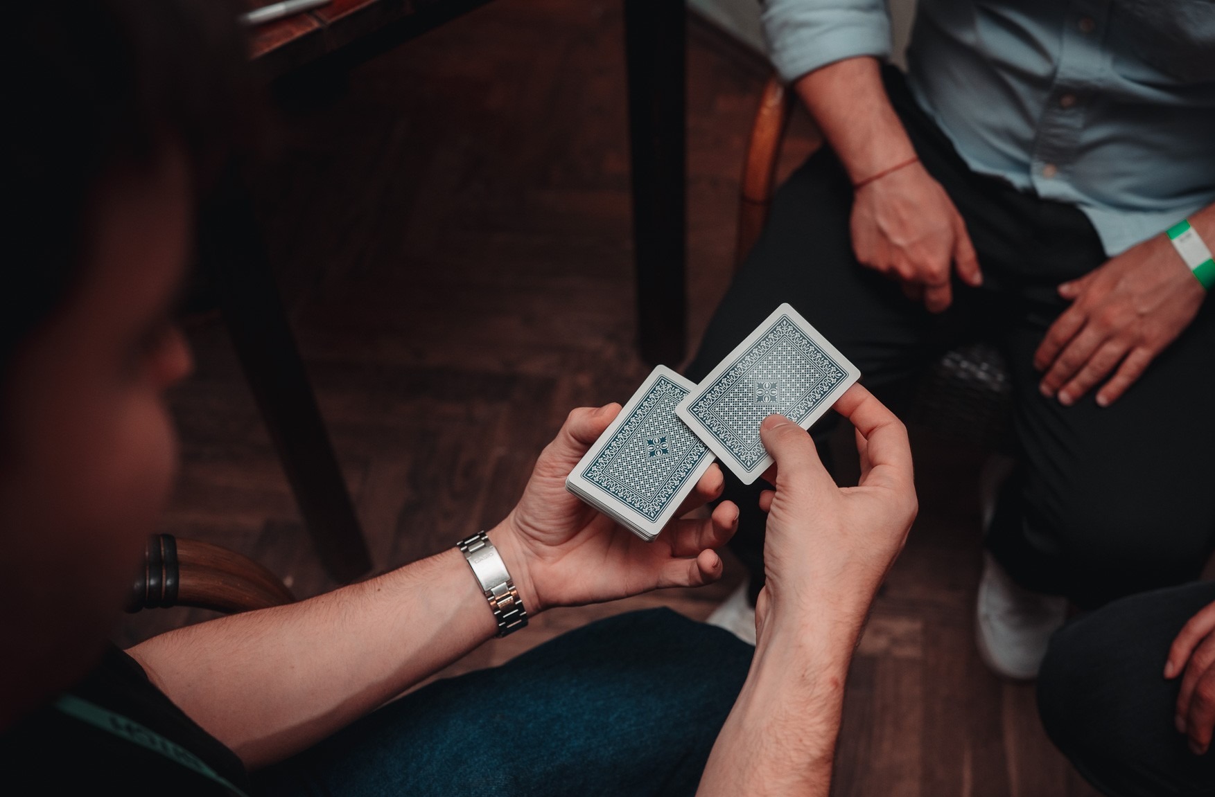 Karty w dłoniach iluzjonisty z Krakowa, przygotowującego się do triku. Widoczny srebrny zegarek na nadgarstku i fragmenty publiczności w tle.