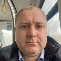 Mężczyzna w kurtce i koszuli w autobusie, widok z bliska na twarz i górną część ciała, za oknem widoczne krople deszczu.