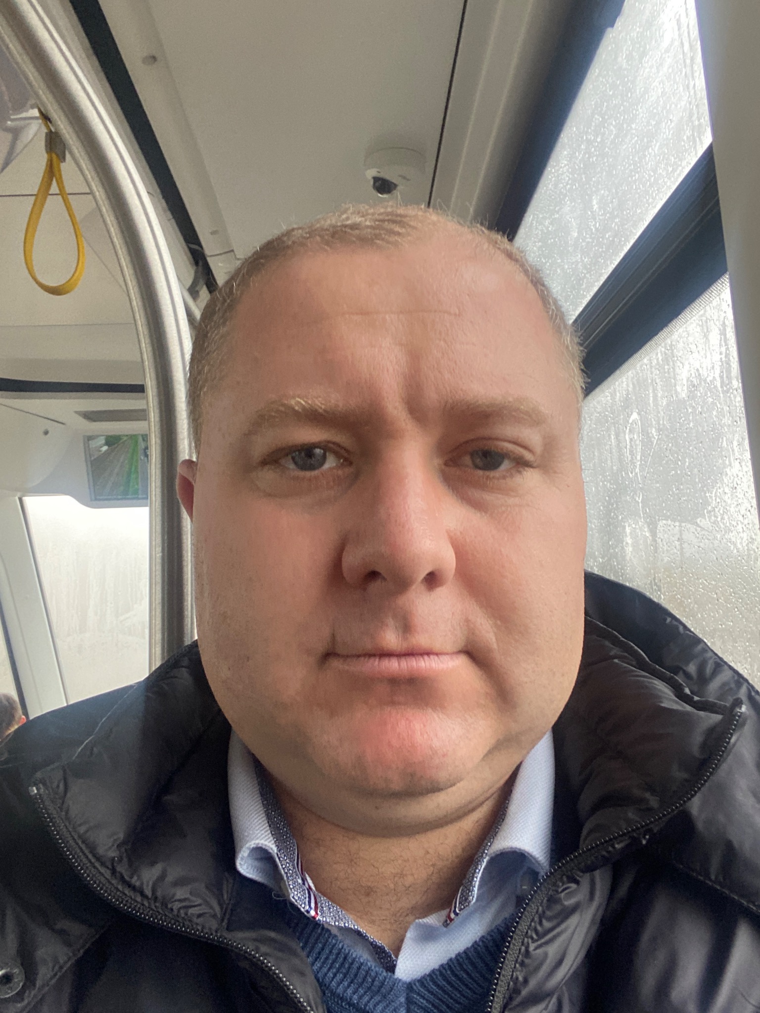Mężczyzna w kurtce i koszuli w autobusie, widok z bliska na twarz i górną część ciała, za oknem widoczne krople deszczu.